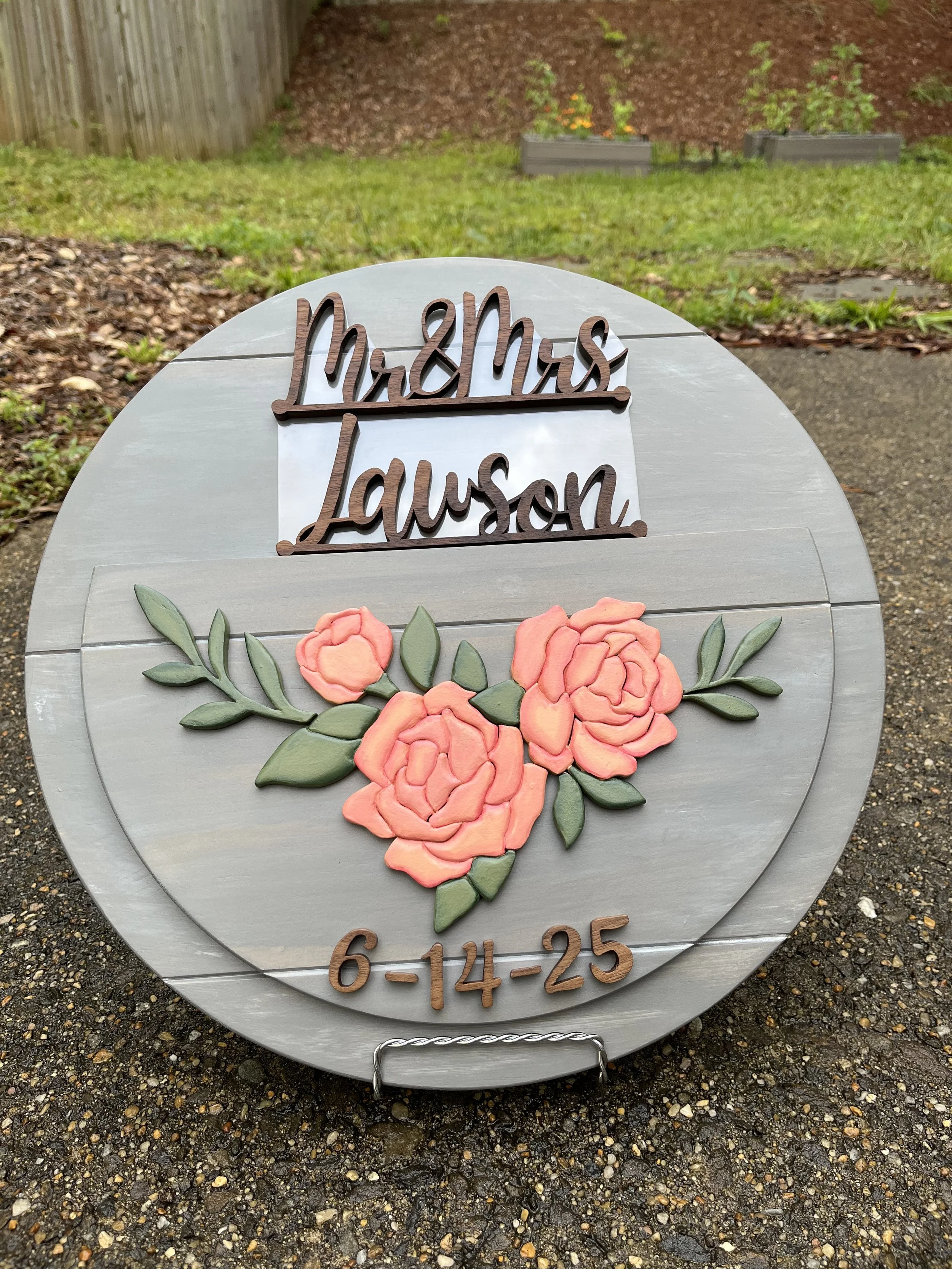 A round wooden wedding sign featuring the text “Mr & Mrs Lawson” in elegant script at the top, with a beautiful arrangement of pink roses and green leaves in the center. The bottom displays the wedding date “6-14-25” in matching script. The sign has 