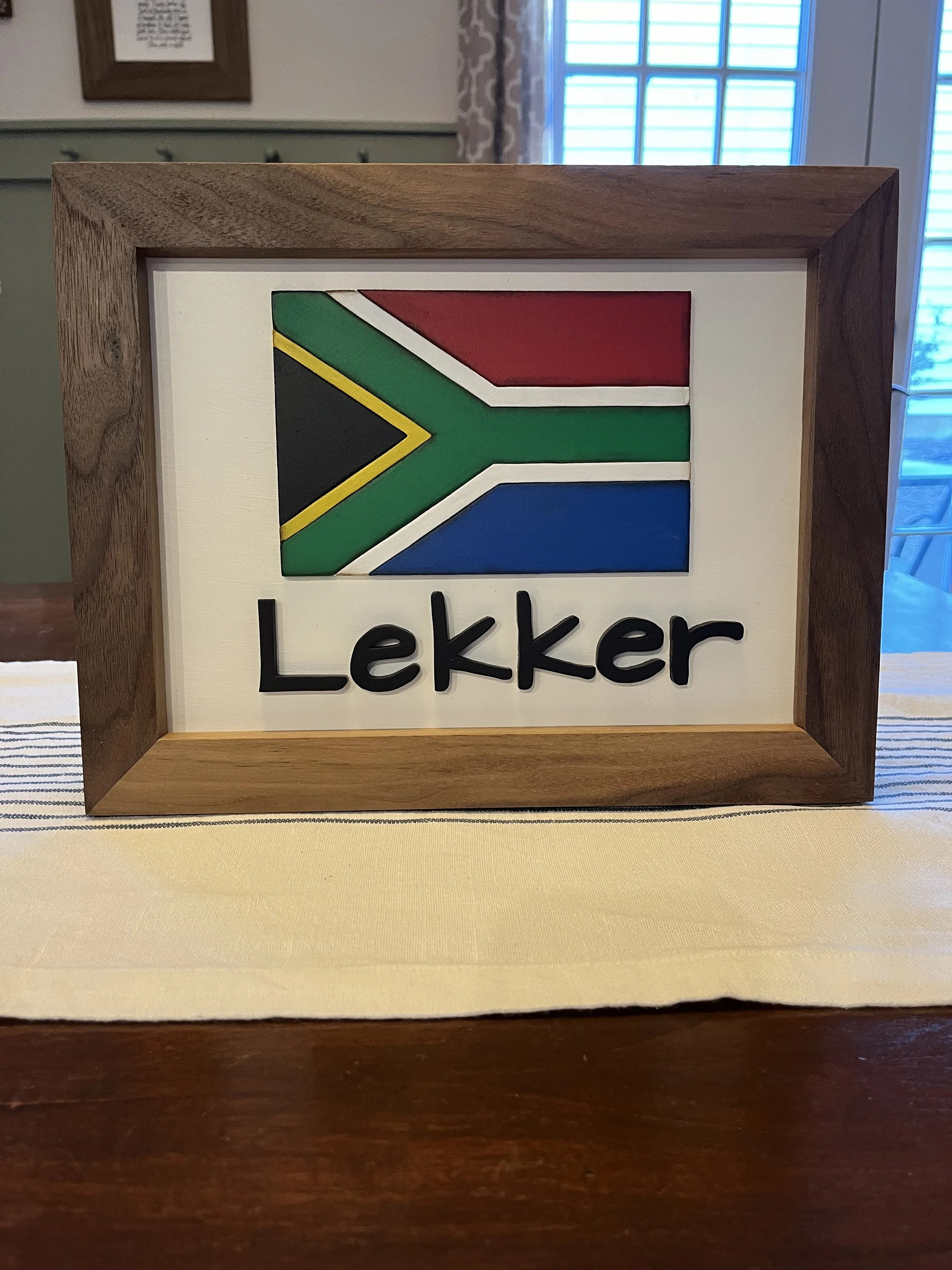 A wooden-framed sign with a background of a white panel, featuring the South African flag above the word "Lekker" in bold black letters.