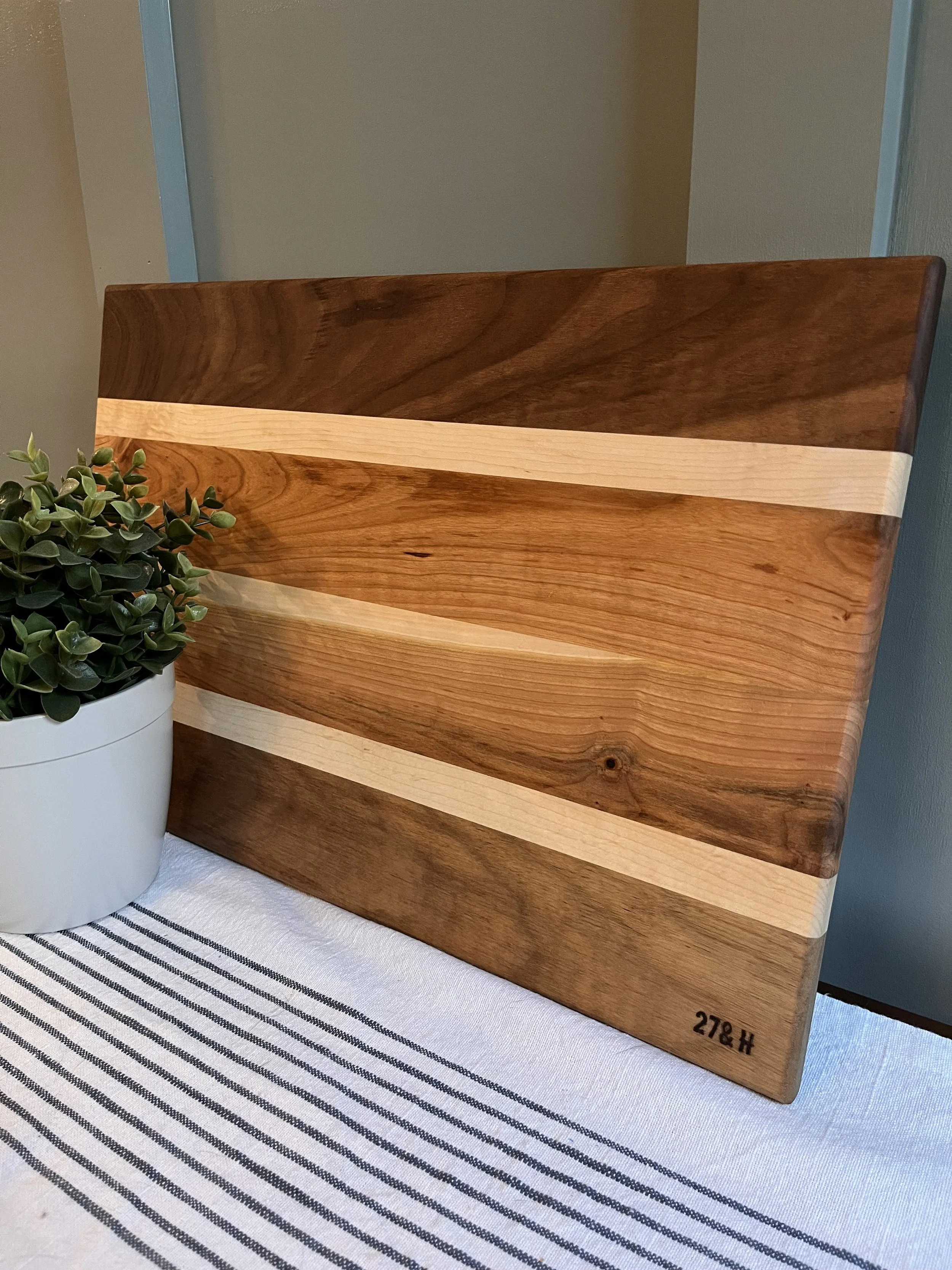A handcrafted wooden cutting board featuring a sleek, modern design with alternating light and dark wood stripes. The board has a smooth finish and natural wood grain patterns, adding a rustic yet elegant touch to any kitchen. A small engraved text “