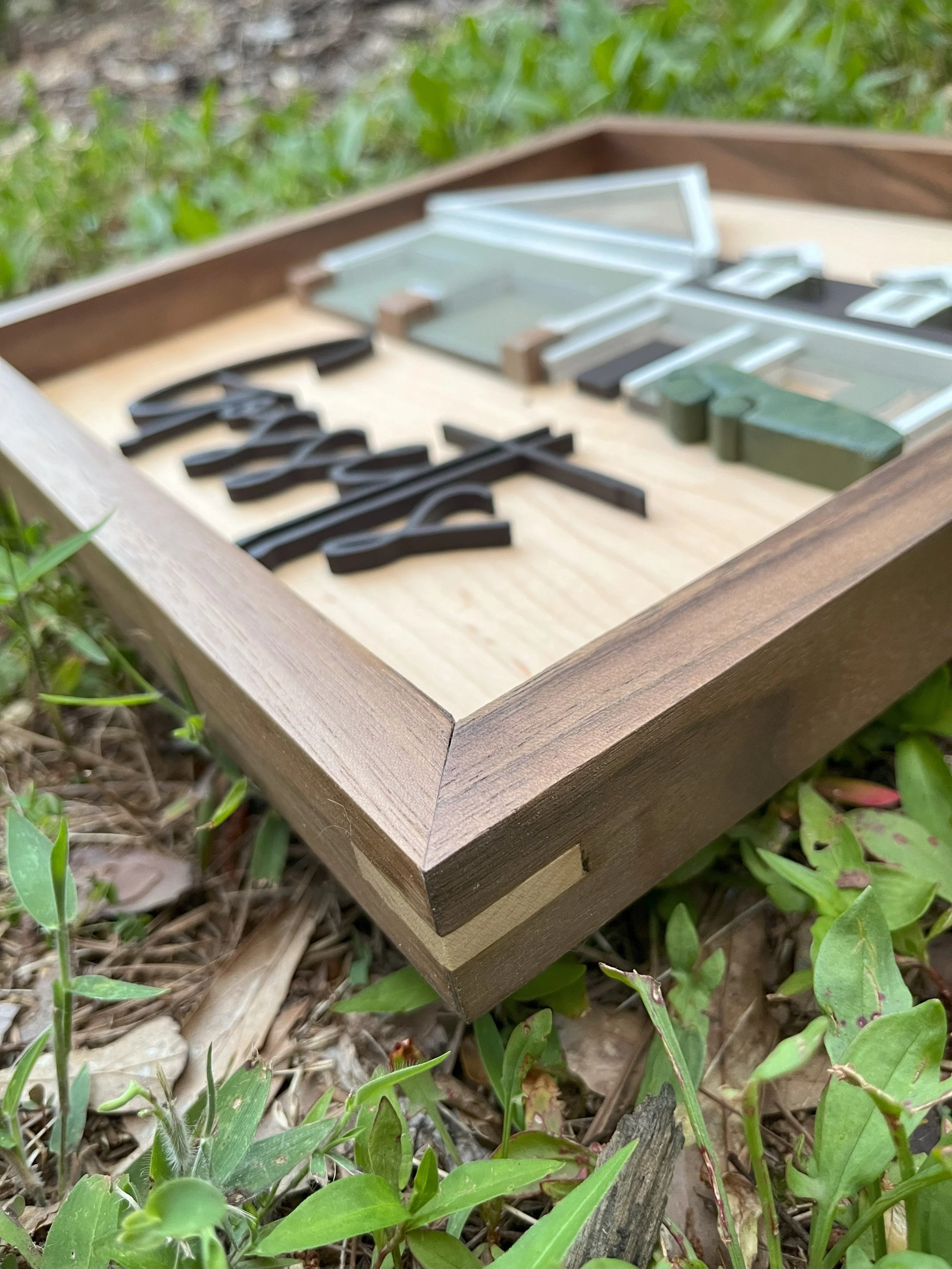 Close-up of a wooden framed sign with black, white, and green decorative characters, resting on green grass and small plants.