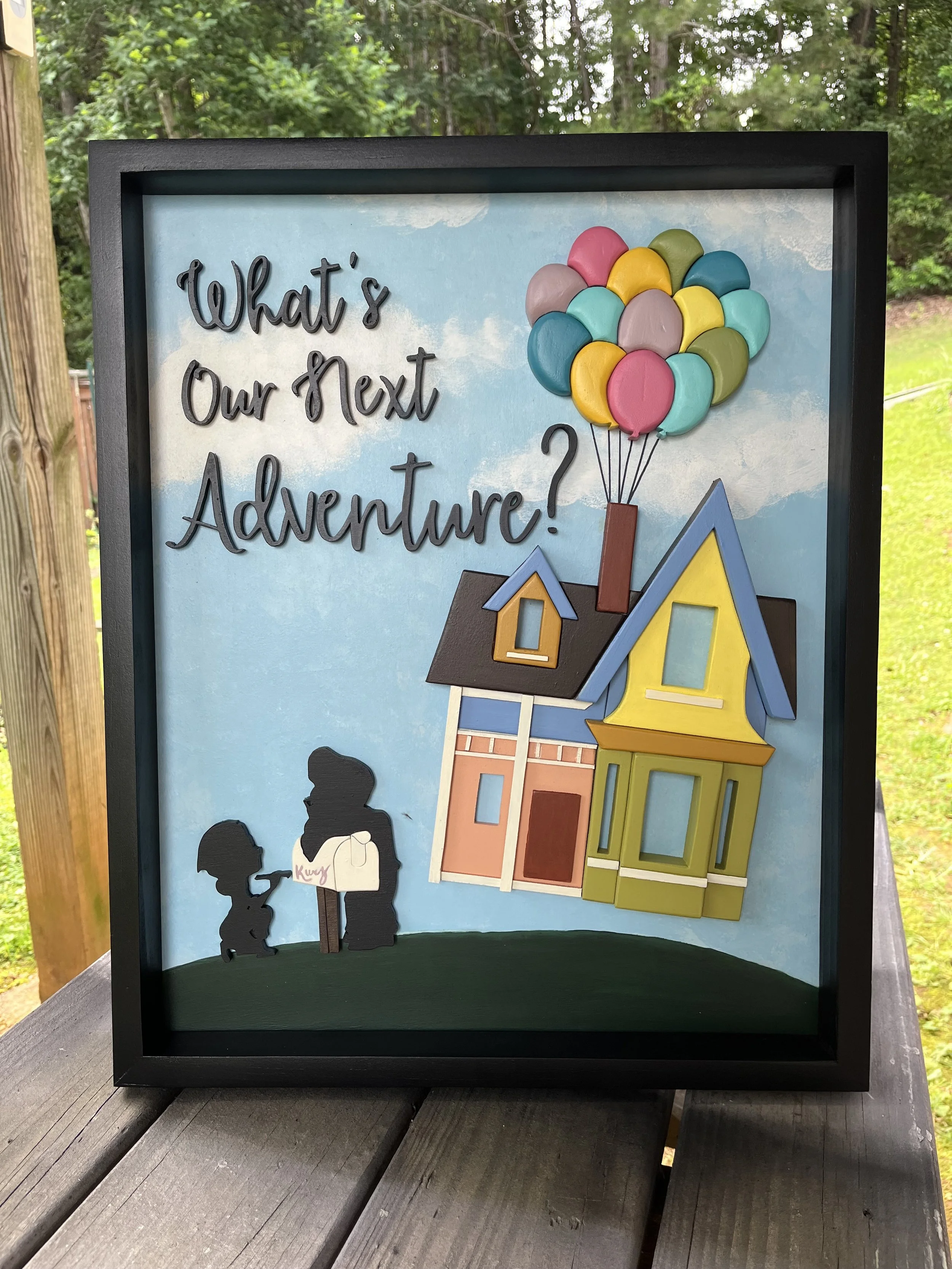 A framed 3D shadow box art piece inspired by the movie Up, featuring a colorful house with balloons floating above it and two silhouetted figures looking at a mailbox. The background includes a bright blue sky with fluffy clouds and the text “What’s 