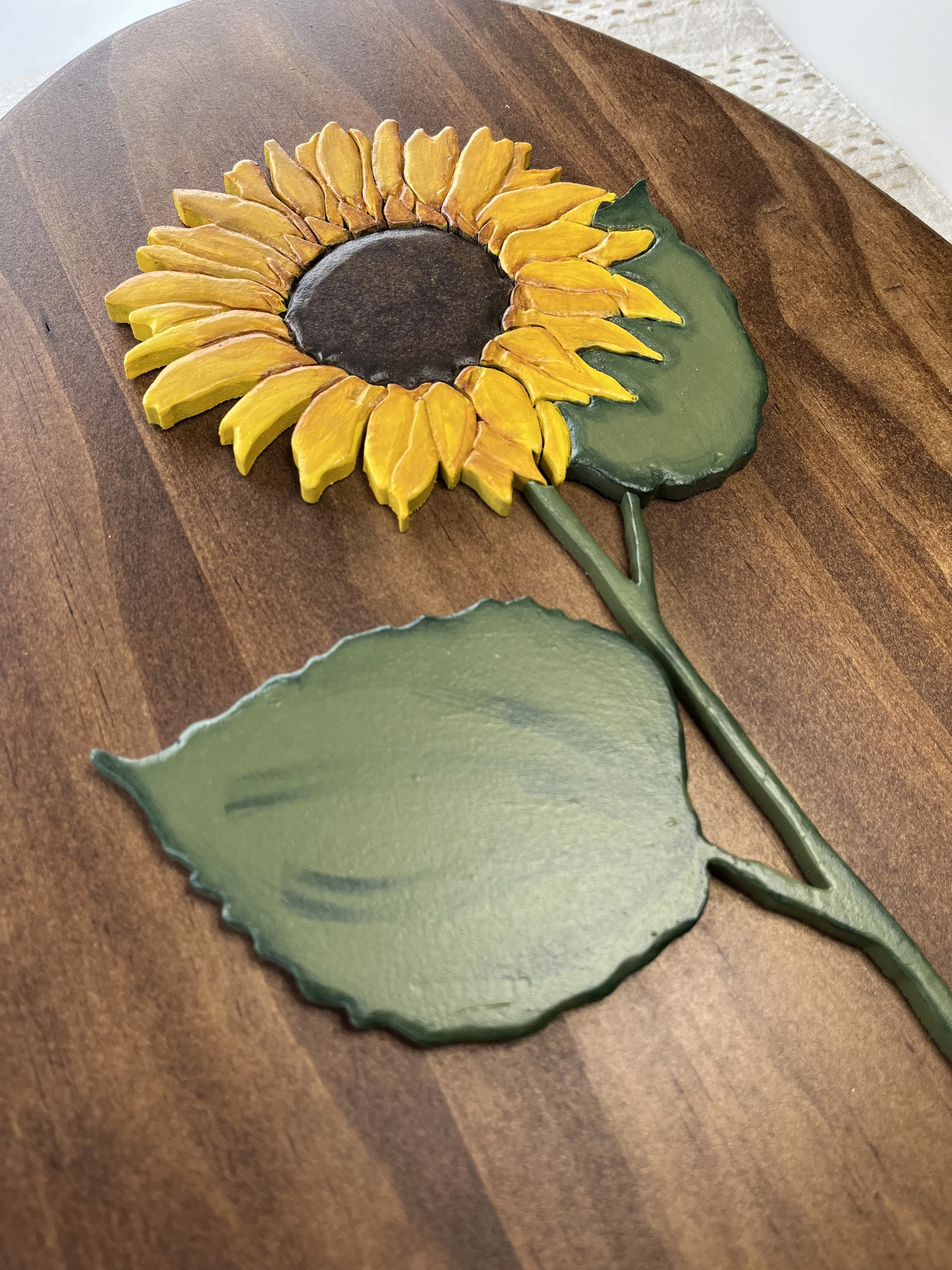 Decorative metal sunflower with yellow petals, a brown center, a green leaf, and a green stem on a wooden surface.