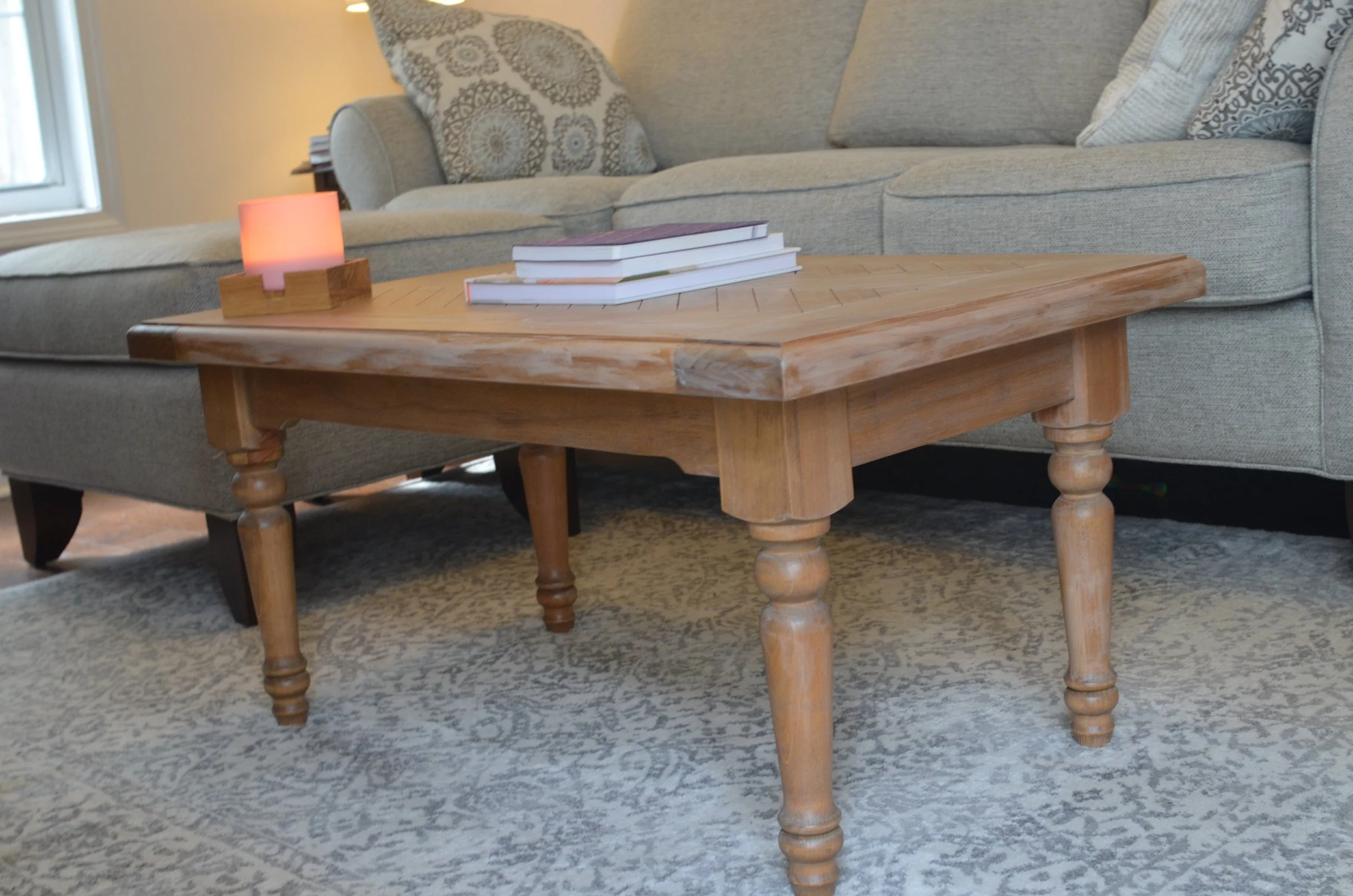 A wooden coffee table with four turned legs in a living room. On top of the table are three stacked books and a lit pink candle in a wooden holder. Behind the table is a gray sofa with patterned pillows, and a light-colored patterned area rug covers 