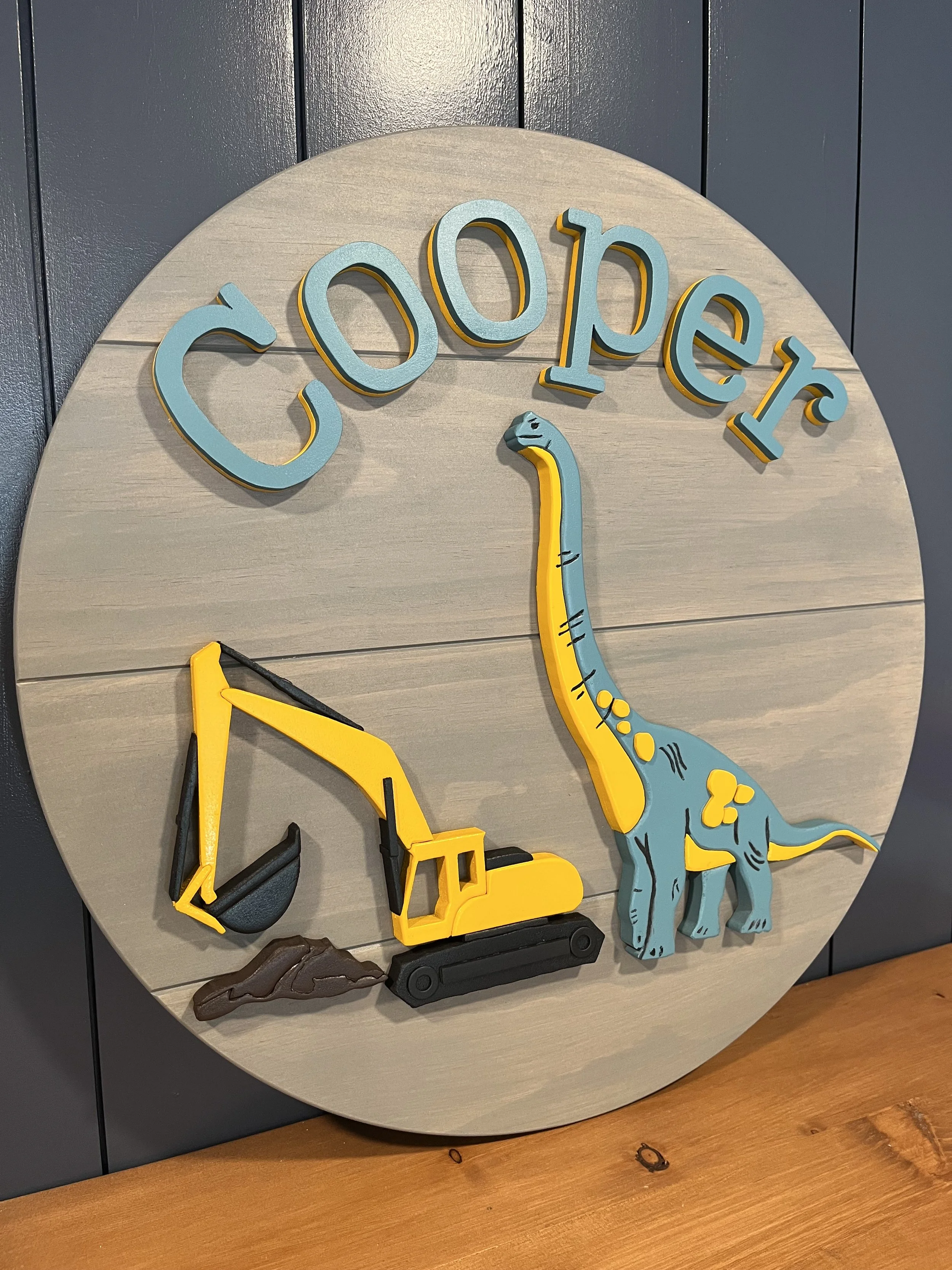 A custom round wooden name sign featuring the name “Cooper” in bold, playful letters at the top. The design includes a bright yellow excavator digging into a small pile of dirt alongside a blue and yellow dinosaur, creating a fun construction and pre