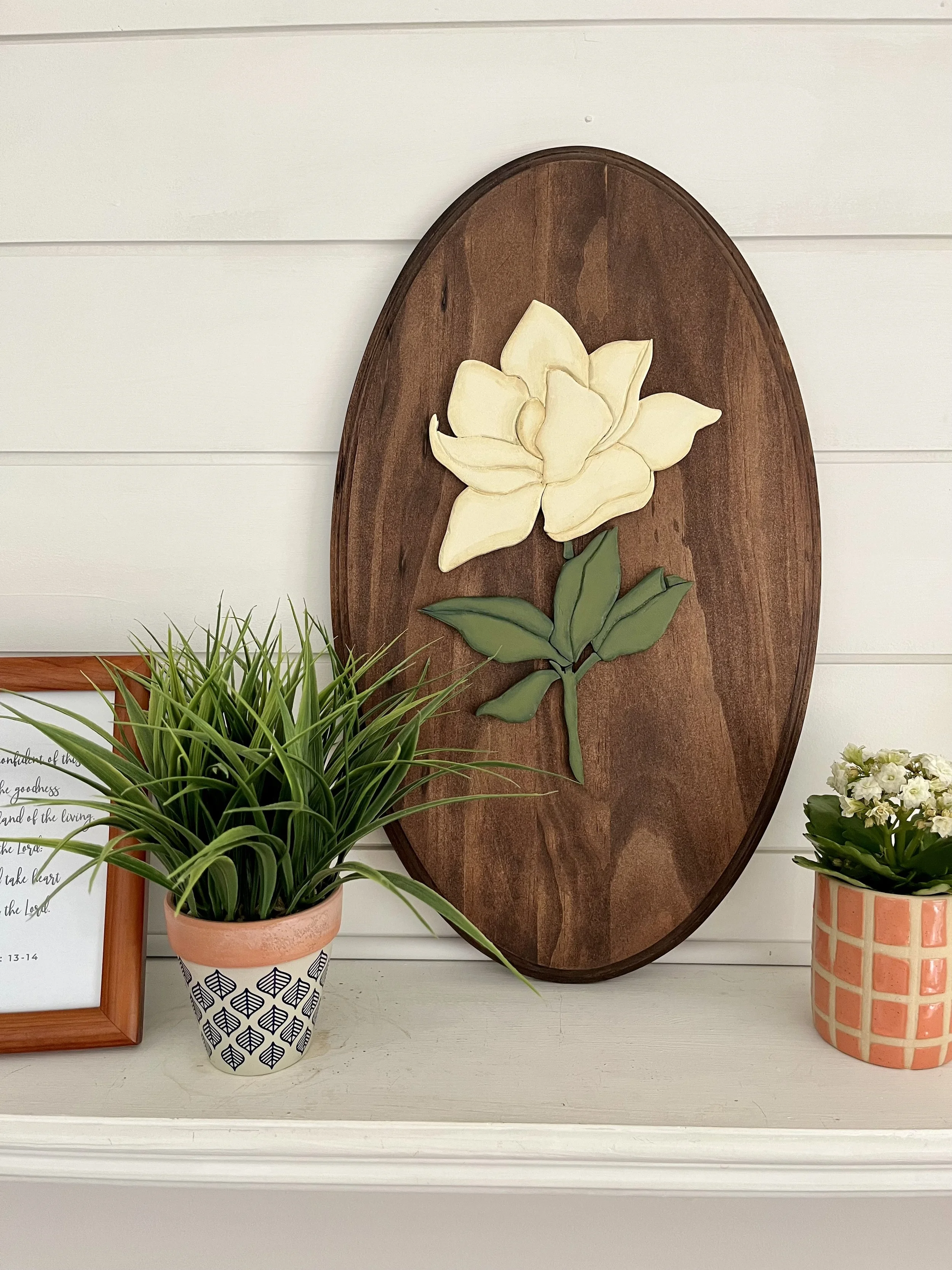Decorative wall art featuring a cream-colored flower on a wooden oval plaque, flanked by two potted plants with green leaves in patterned pots, on a white surface in front of white shiplap wall.