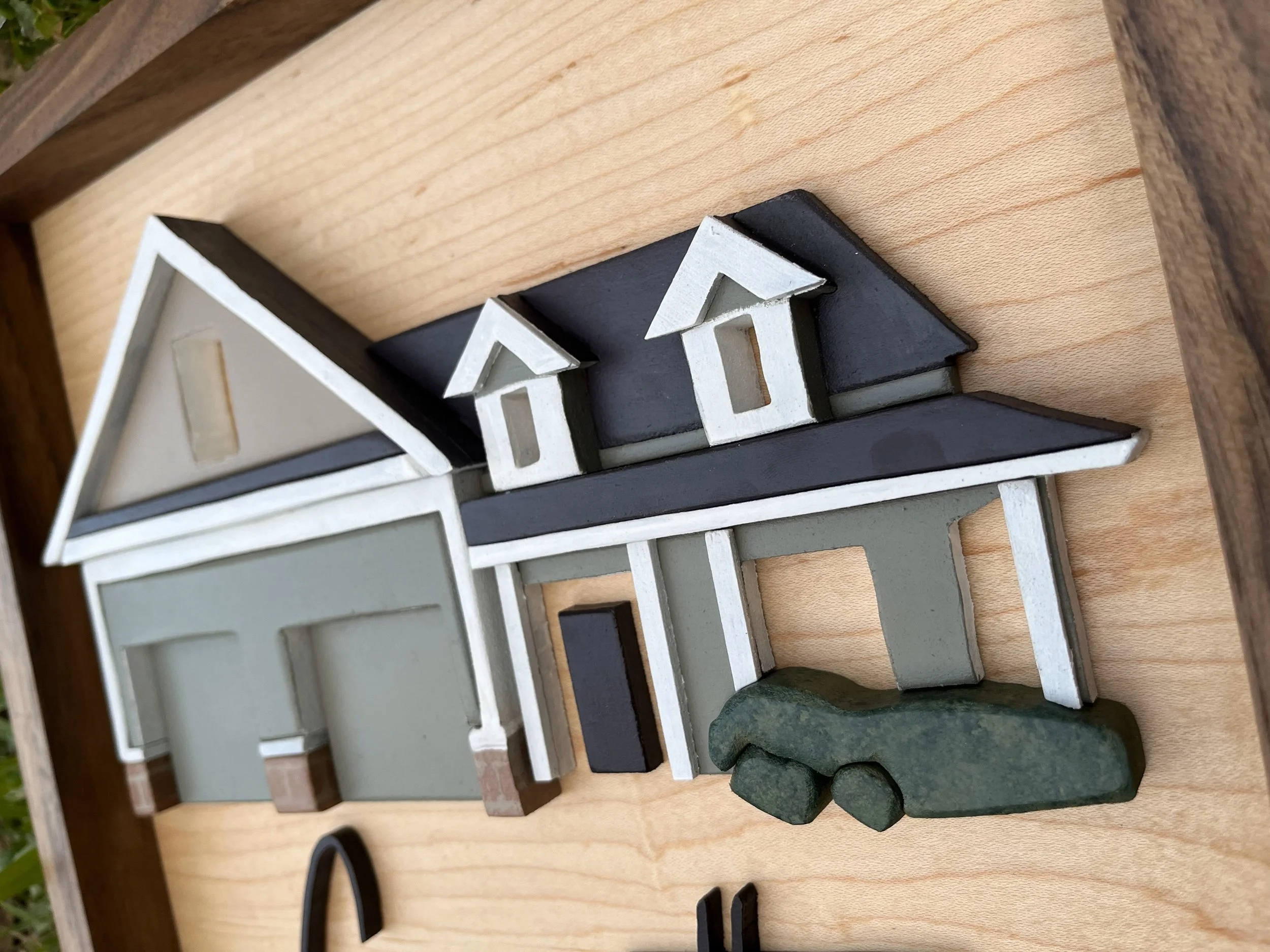 Wooden decorative piece depicting a house with a dark roof, two small dormer windows, and a fenced area, placed on a wooden surface.