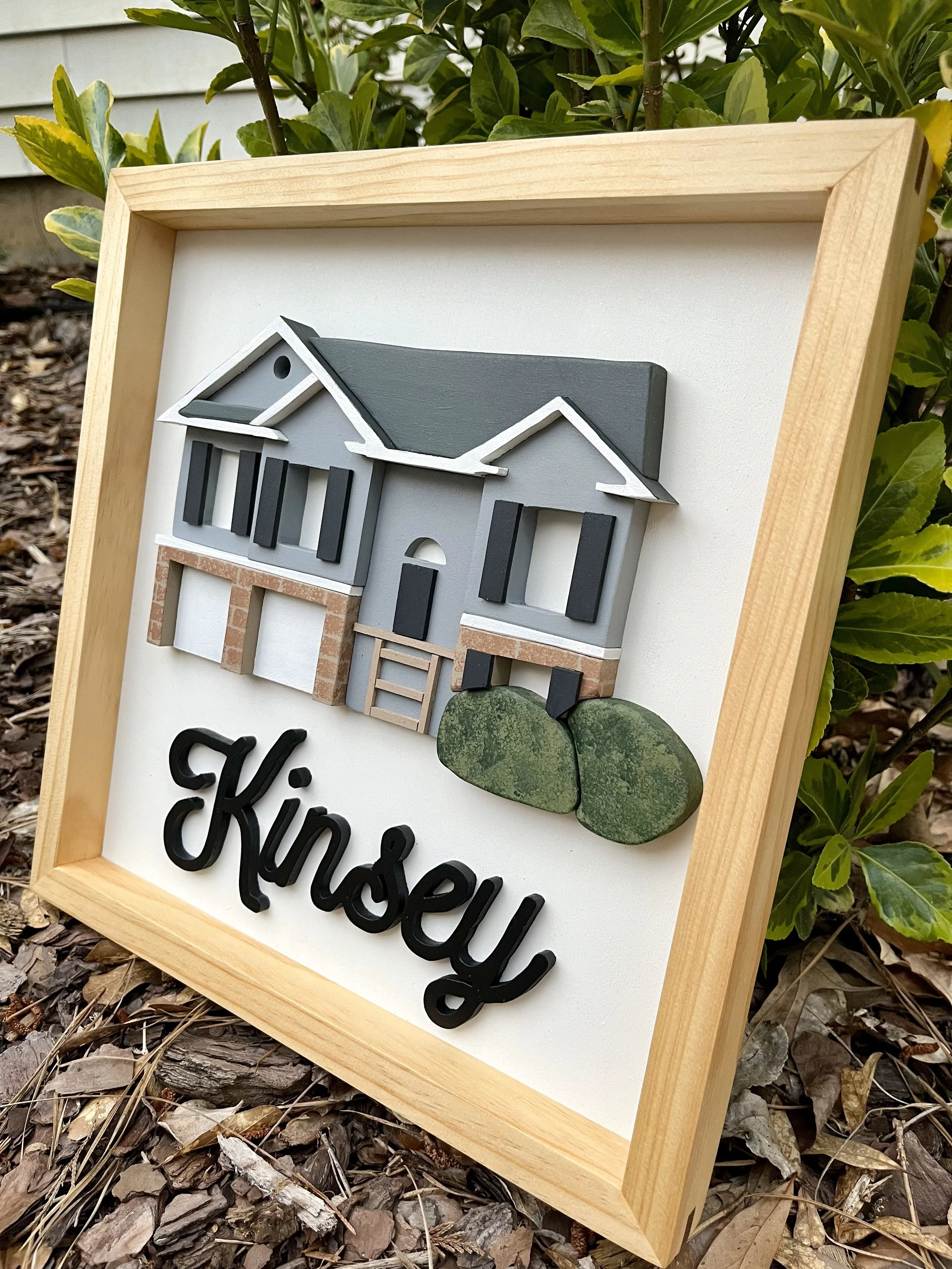 Decorative house-shaped wall sign with the word 'Kinsley' in black cursive letters below a model of a house with gray and white details, brick accents, and a small green bush at the bottom.