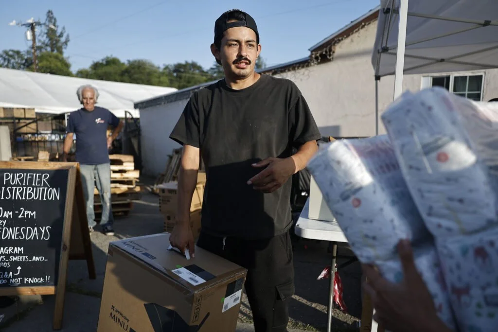 A young man in a black t-shirt and backwards cap handing a package to a person at an outdoor event. A man stands in the background near a blackboard sign, under a white tent.