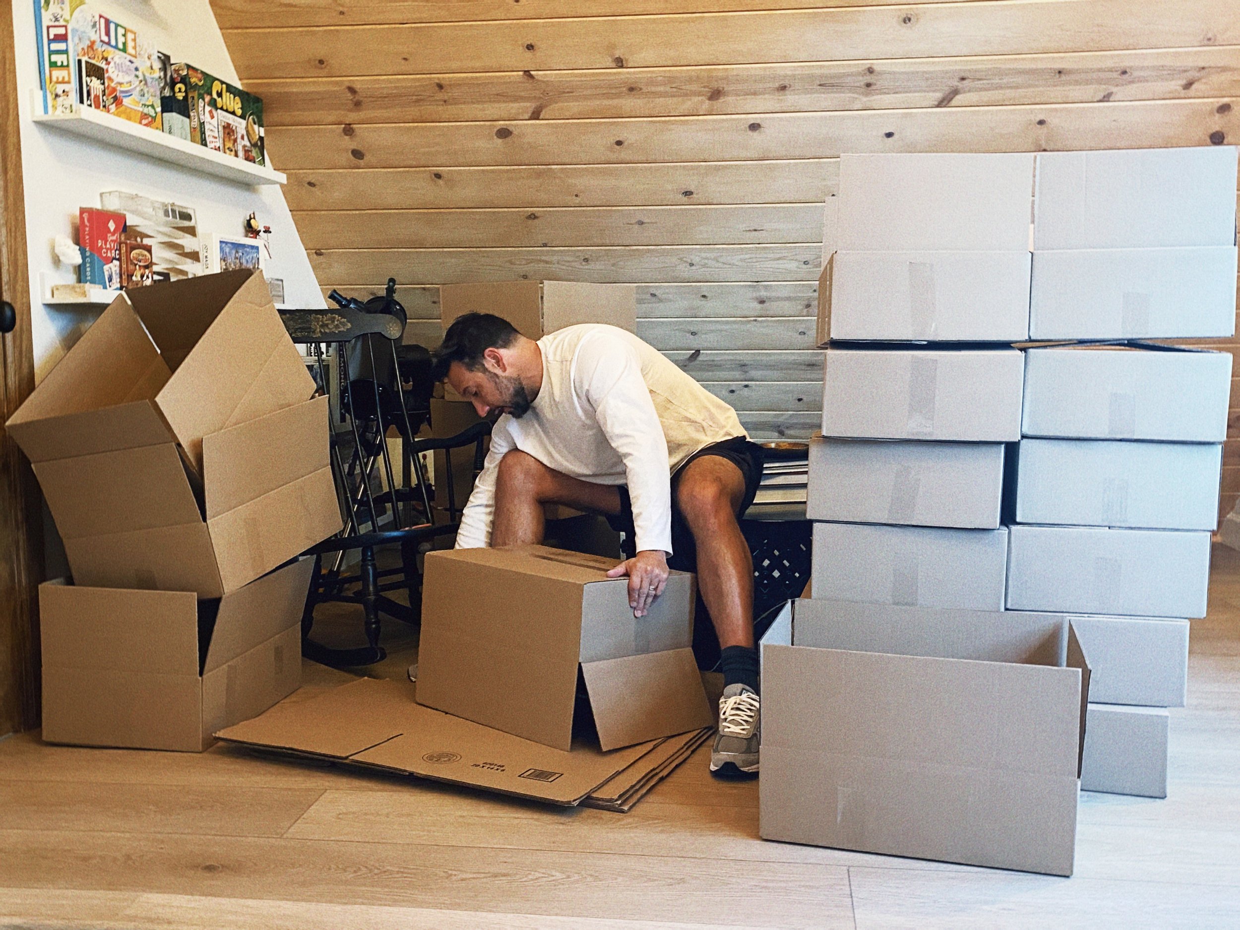 Man filling boxes with items in a room with a wooden wall, surrounded by stacked cardboard boxes and shelves of board games.