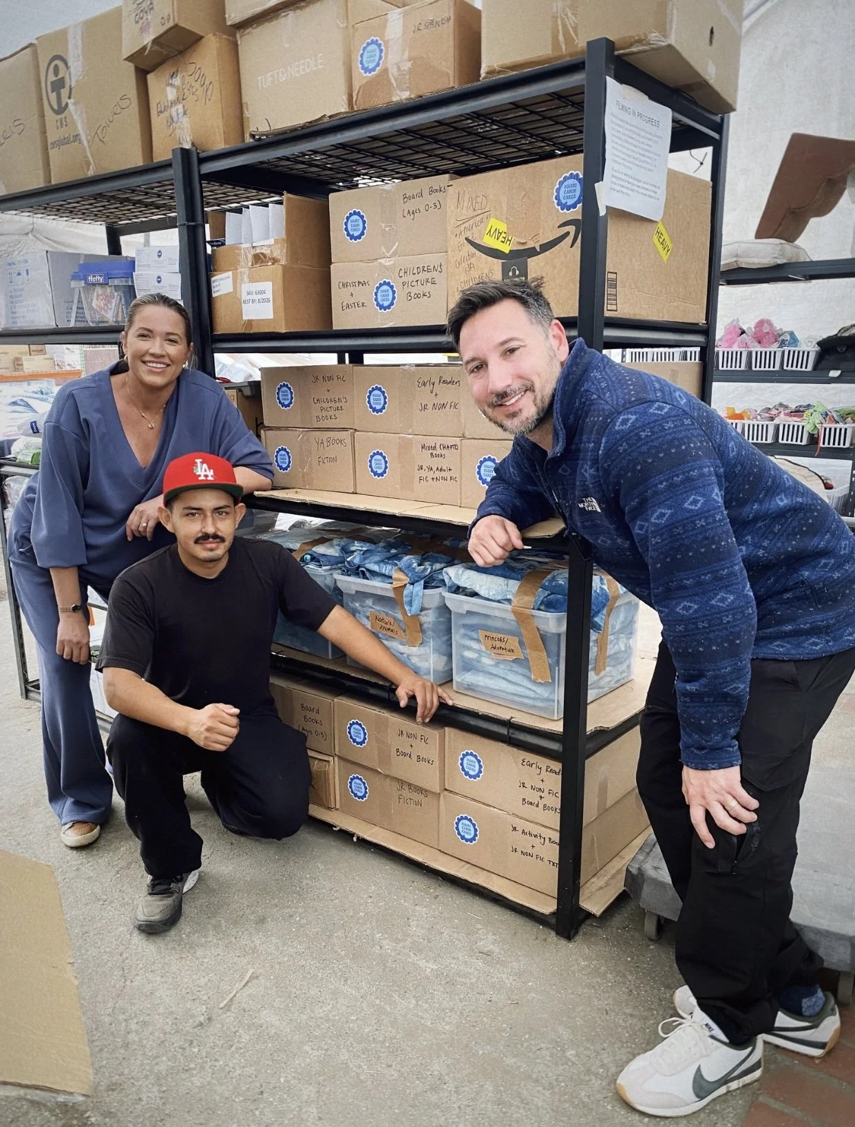 Three people posing in front of a metal shelf filled with cardboard boxes containing donated books, likely in a warehouse or donation center.
