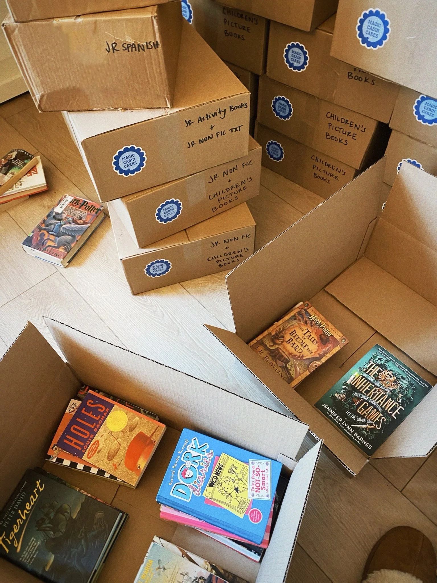 Several cardboard boxes labeled with handwritten notes such as 'Jr Spanish,' 'Jr Activity Books,' 'Jr Non-Fic,' and 'Children's Picture Books,' placed on a wooden floor. Some boxes contain children's and young adult books, including Harry Potter and other novels, with a few books laid out on top of the boxes.