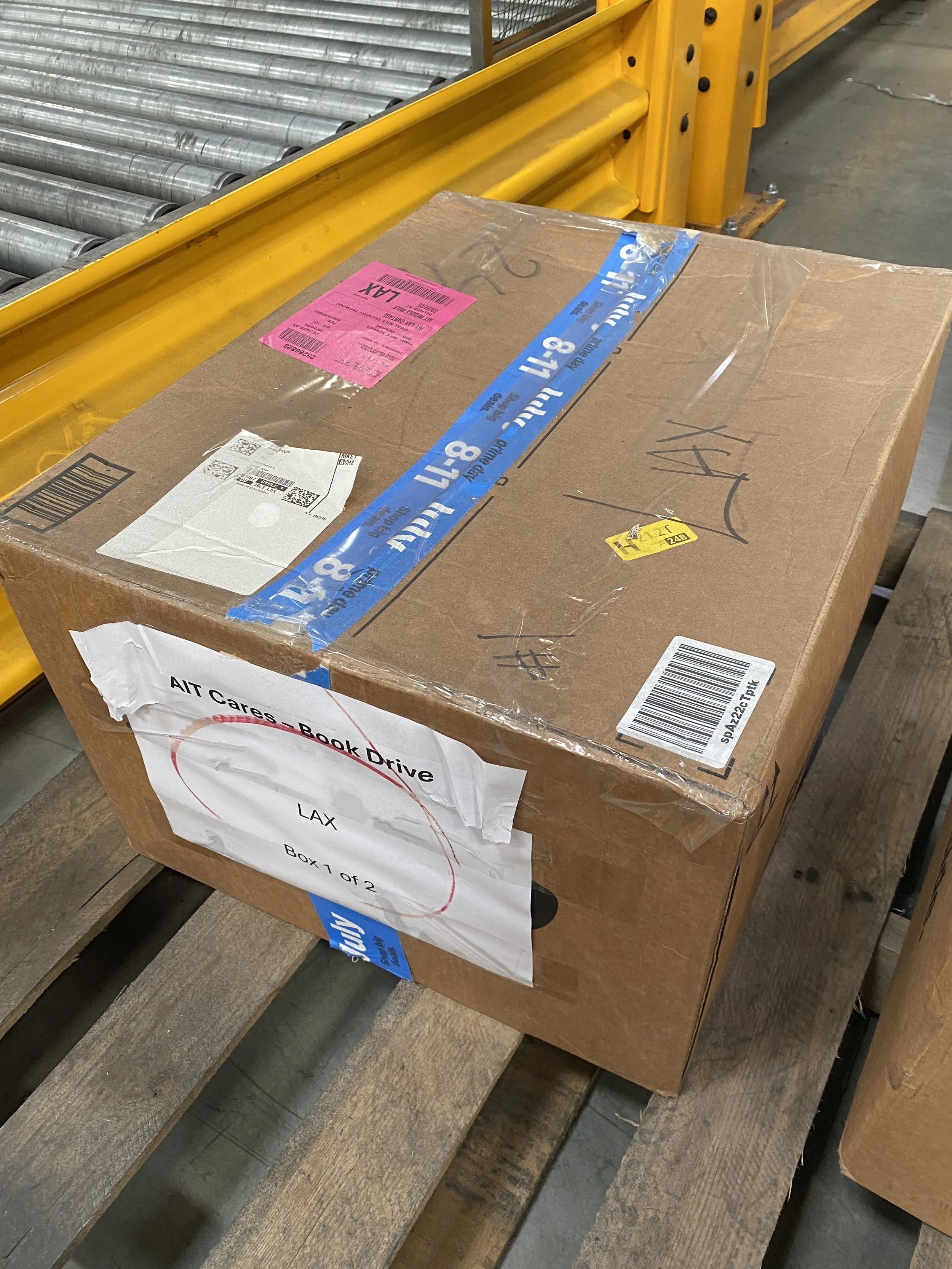 A cardboard box labeled 'AIT Cares - Book Drive' with additional labels including 'LAX' and 'Box 1 of 2', secured with blue tape, placed on a wooden pallet in a warehouse.