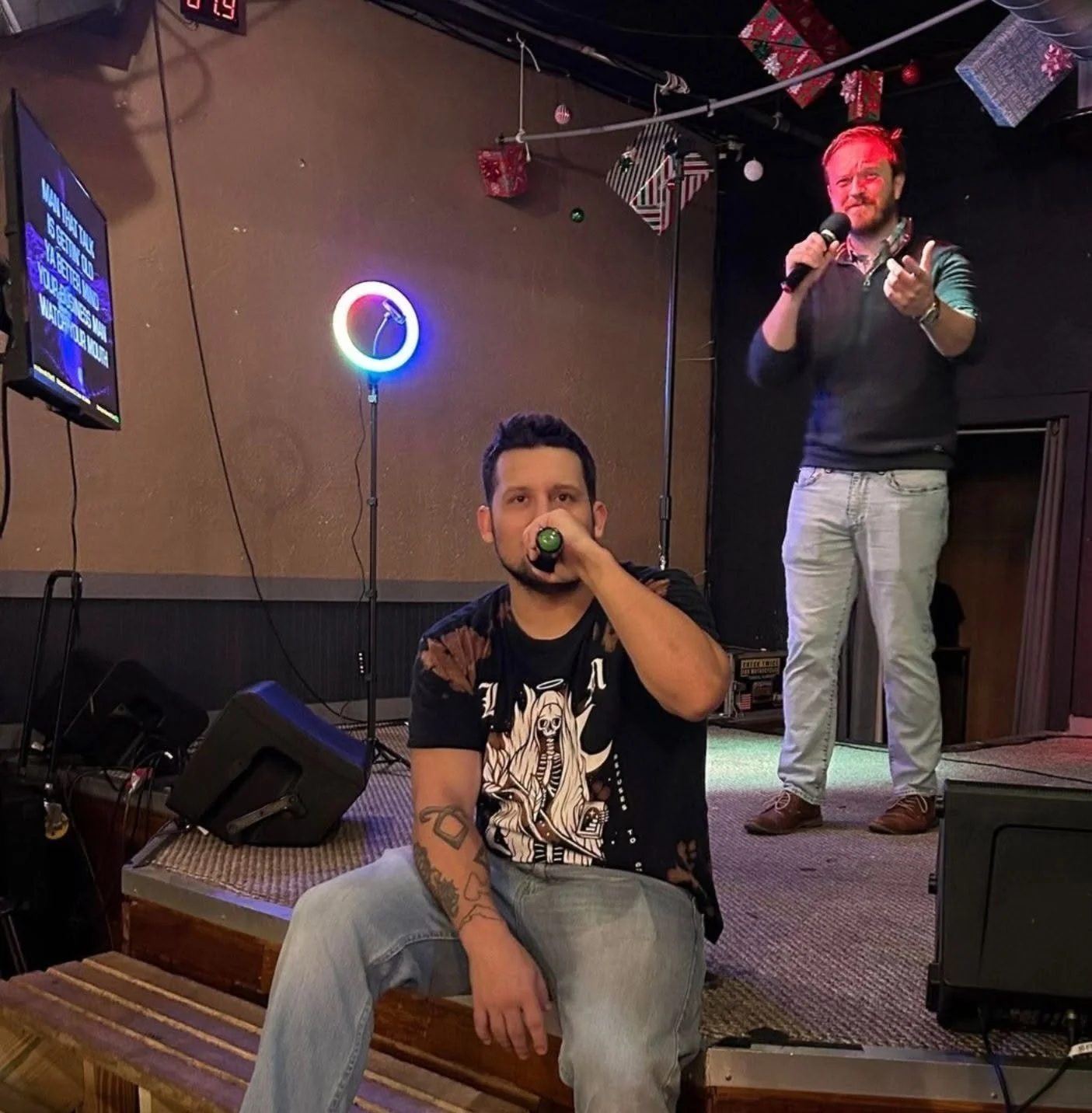 Two men performing karaoke in a dimly lit venue decorated for Christmas, with one sitting on a bench and singing into a microphone, and the other standing on stage with a microphone in hand, gesturing towards the audience.