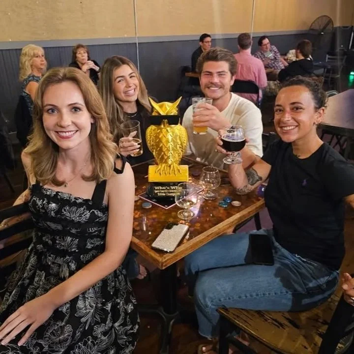 Four friends sitting at a table in a bar or restaurant, smiling at the camera, with drinks in hand and a large yellow pineapple-shaped trophy in the center.