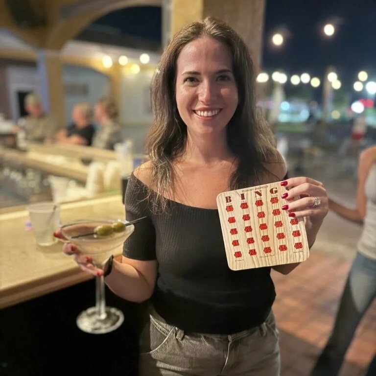 A woman with long brown hair smiling inside a bar or restaurant, holding a cocktail glass with a drink and a garnish in her left hand, and a wooden bingo card in her right hand. The background shows other people sitting and socializing.