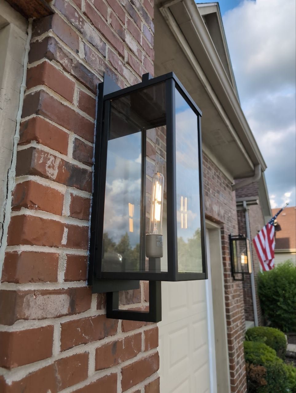 Close-up of a black outdoor wall-mounted lantern with a glass enclosure, attached to a brick wall outside a house, with a cloudy sky reflected in the glass.
