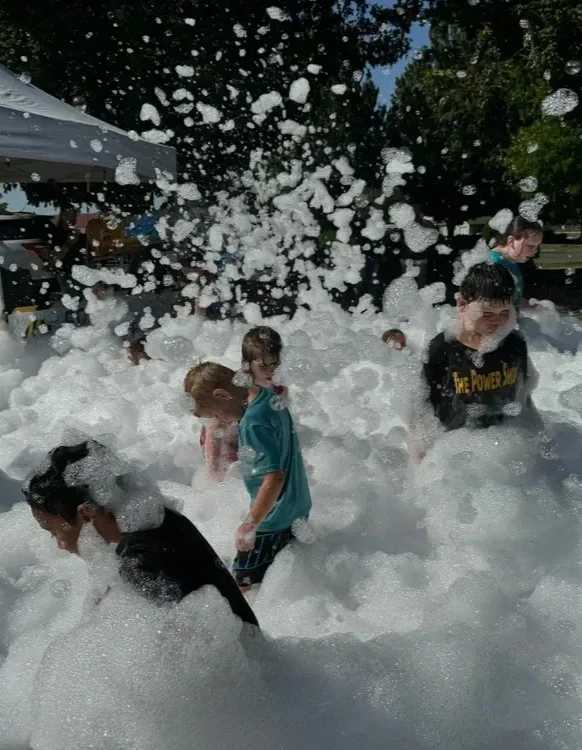 Children playing in a foam party with large amounts of foam and bubbles outdoors on a sunny day.
