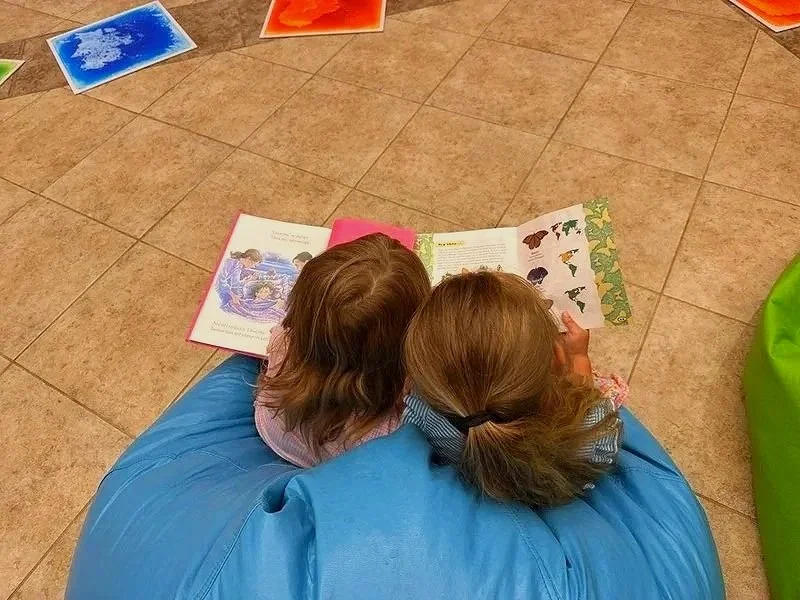 Two young children sitting on a blue bean bag chair, reading a colorful picture book on a tiled floor.