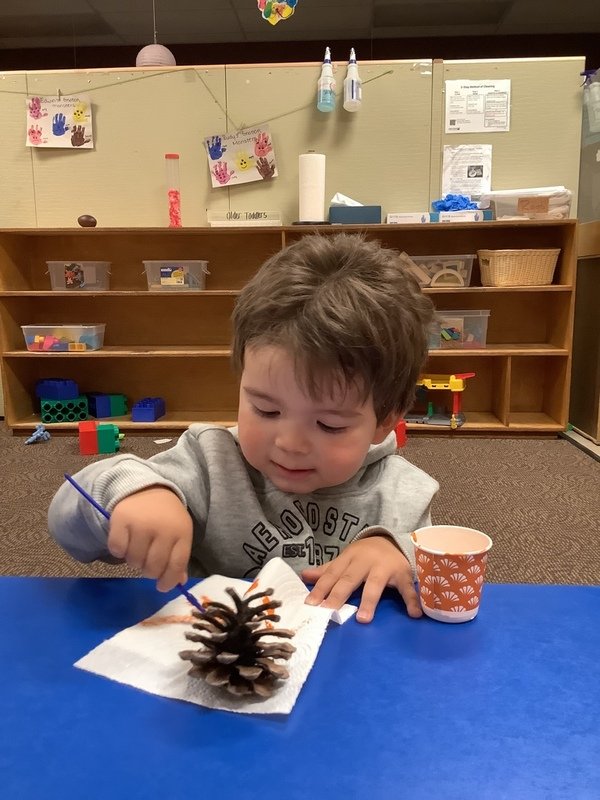 A young boy with brown hair, wearing a gray sweatshirt, is sitting at a blue table in a classroom. He is using a paintbrush to decorate a pinecone placed on a paper towel. There is an orange paper cup beside him. In the background, there are shelves with toy building blocks, craft supplies, and children's artwork on the wall.