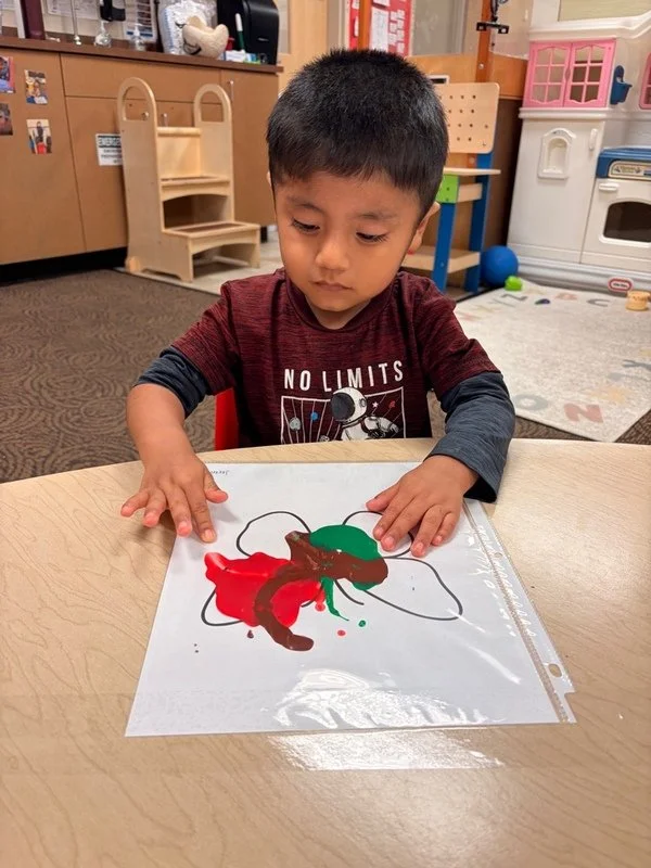 Young boy in a red shirt seated at a table, painting on a paper with red, green, and brown paint in a classroom setting.
