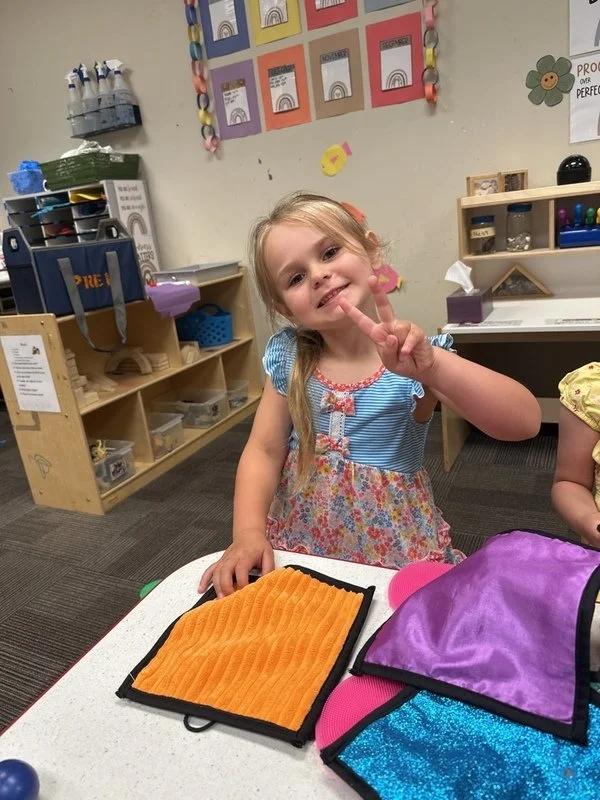 A young girl with blonde hair in pigtails smiling and making a peace sign with her fingers in a classroom.