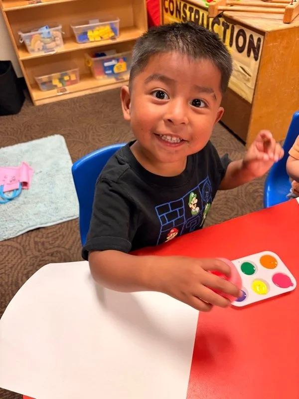 A young boy smiling and sitting at a red and white table in a classroom, playing with a color match game featuring six colorful circles.