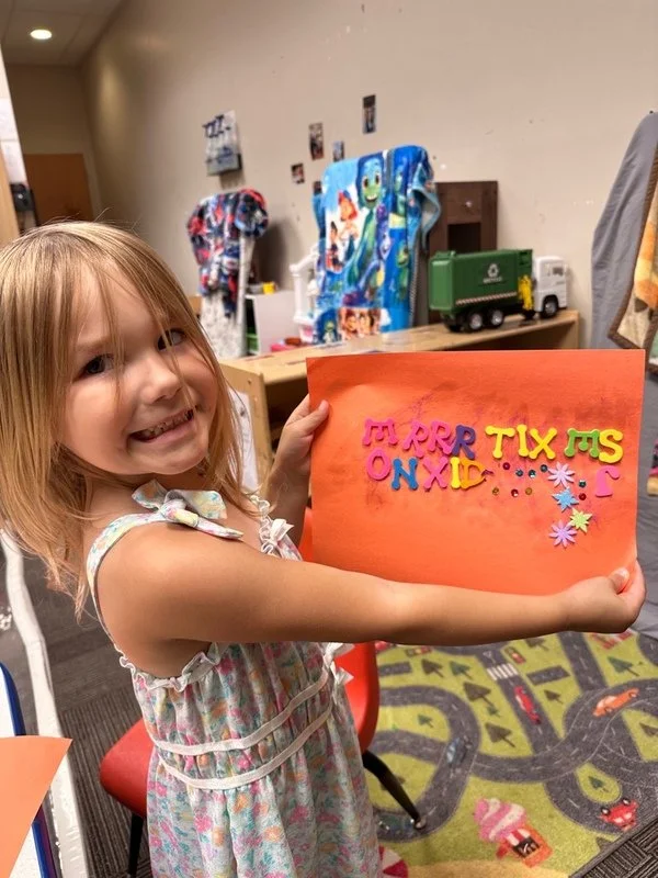 A young girl with red hair, wearing a colorful dress, is smiling and holding a bright orange paper with multicolored foam letters and decorations. The background features a playroom with blankets, a toy truck, and a colorful play mat with roads and cars.