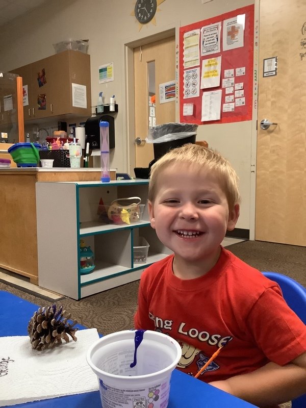 A young boy with blonde hair and a red T-shirt smiling at a classroom table. On the table, there's a pine cone and a cup with some blue and orange string inside. The classroom has educational posters on the wall, a counter with supplies, and a clock showing 3:10.