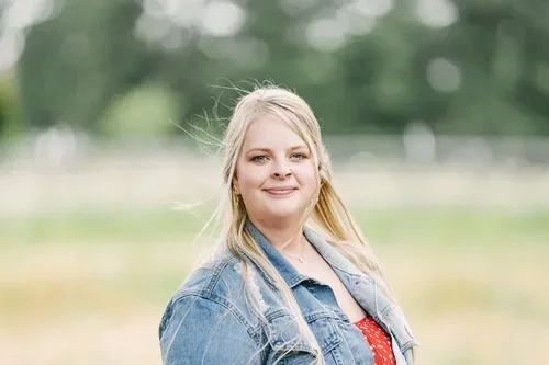 A young woman with long blonde hair smiling outdoors in a park, wearing a denim jacket and red top.