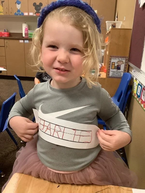 A young girl with curly blonde hair, wearing a gray Nike shirt, a purple headband, and a light brown tutu, sitting at a classroom table. She is holding a paper with the name 'HARLEY' written on it, positioned across her chest.