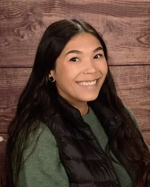 A young woman with long dark hair, smiling, wearing a black vest and green sweater, standing in front of a wooden background.
