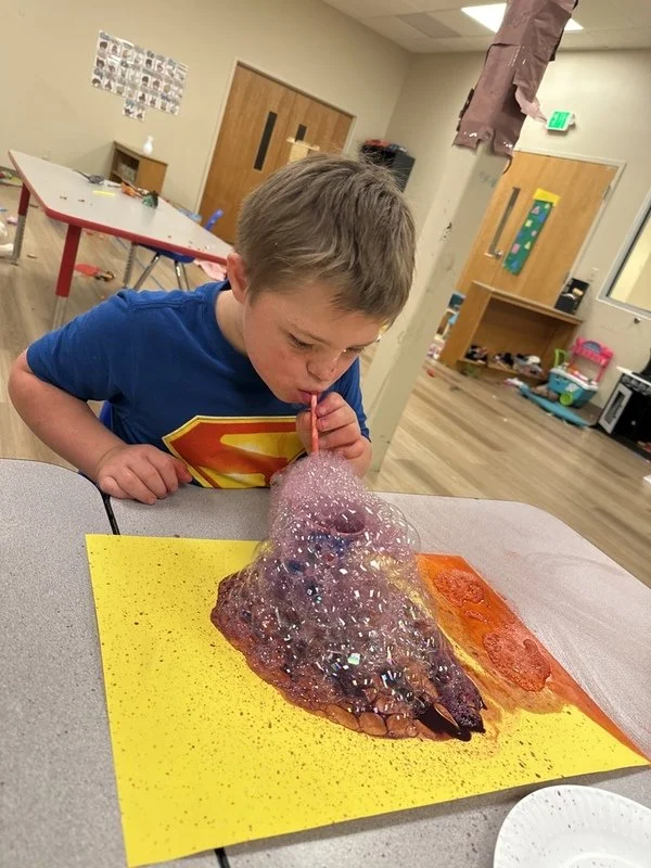 A young boy dressed in a Superman T-shirt blows a large bubble that covers a model of a dinosaur on a table in a classroom.