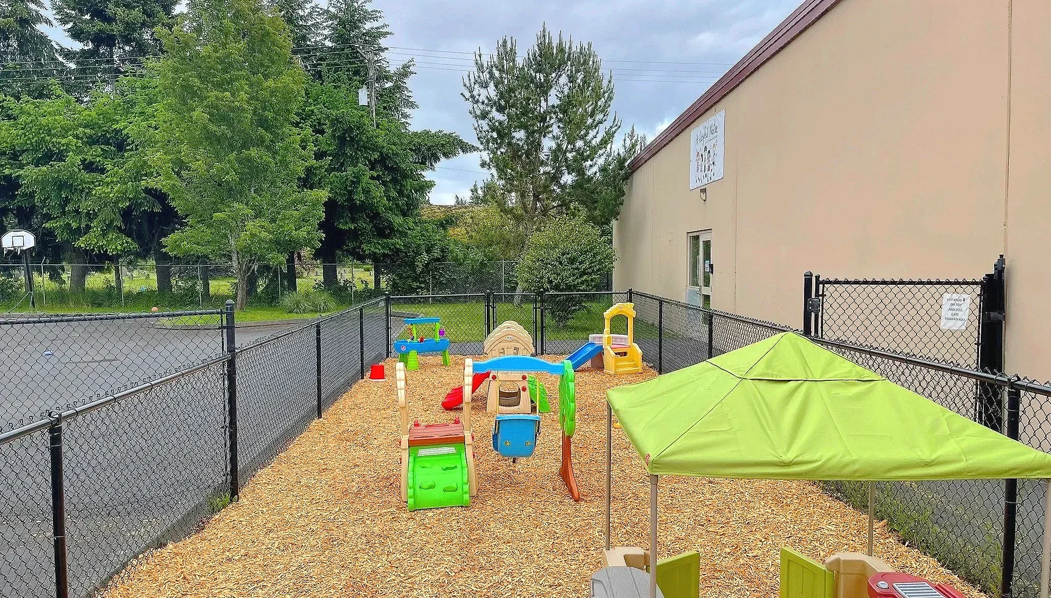 An outdoor children’s play area enclosed by black chain-link fencing. Inside, there are small colorful plastic play structures, a green tent, and a gravel surface. In the background, there are trees, a beige building with a sign, and a cloudy sky.