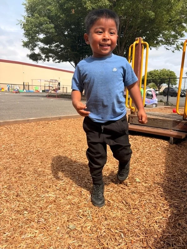 A young boy smiling and running at a playground, wearing a blue T-shirt and black pants, with a play structure and trees in the background.