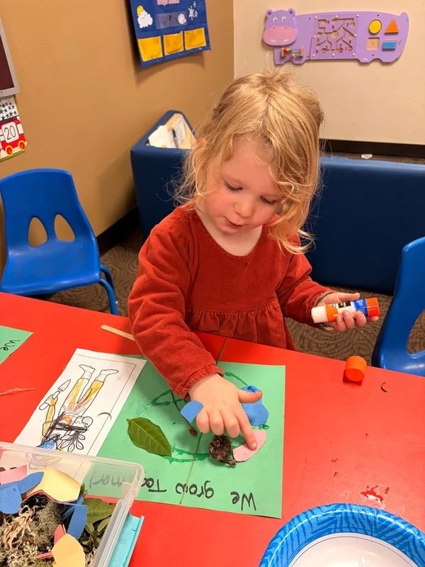 A young girl with curly blonde hair wearing a red long-sleeve dress is sitting at a red table, engaged in a craft project with glue, leaves, and a black material on a green paper.
