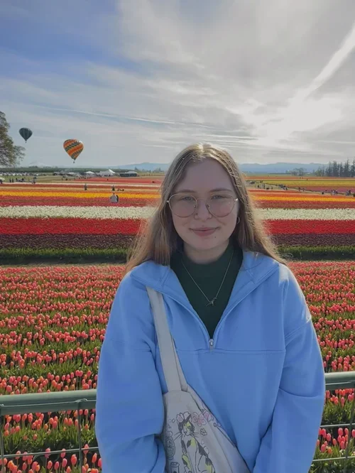 A young woman with glasses standing in front of a colorful tulip field under a partly cloudy sky with hot air balloons in the background.