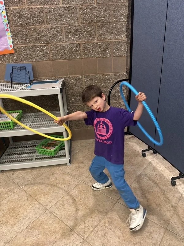 A young boy playing with two hula hoops, one yellow and one blue, in a room with a brick and blue wall, a metal shelf with green baskets, and a tiled floor.