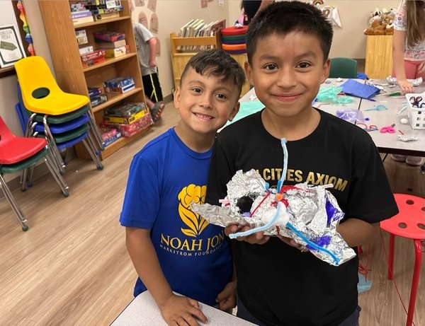 Two boys smiling, one holding a handmade craft with a candle on top in a classroom setting.