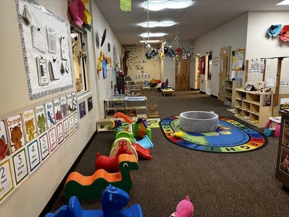 An indoor preschool or daycare classroom decorated with colorful children's artwork, educational posters, and various toys including animal-shaped ride-on toys. There is a circular rug with a clock and alphabet design and a large gray circular reading or activity area in the room, which is well-lit and organized.