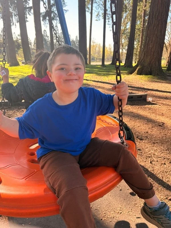 A young boy in a blue shirt and brown pants smiling while sitting on an orange swing at a park, with trees and another child on a swing in the background.