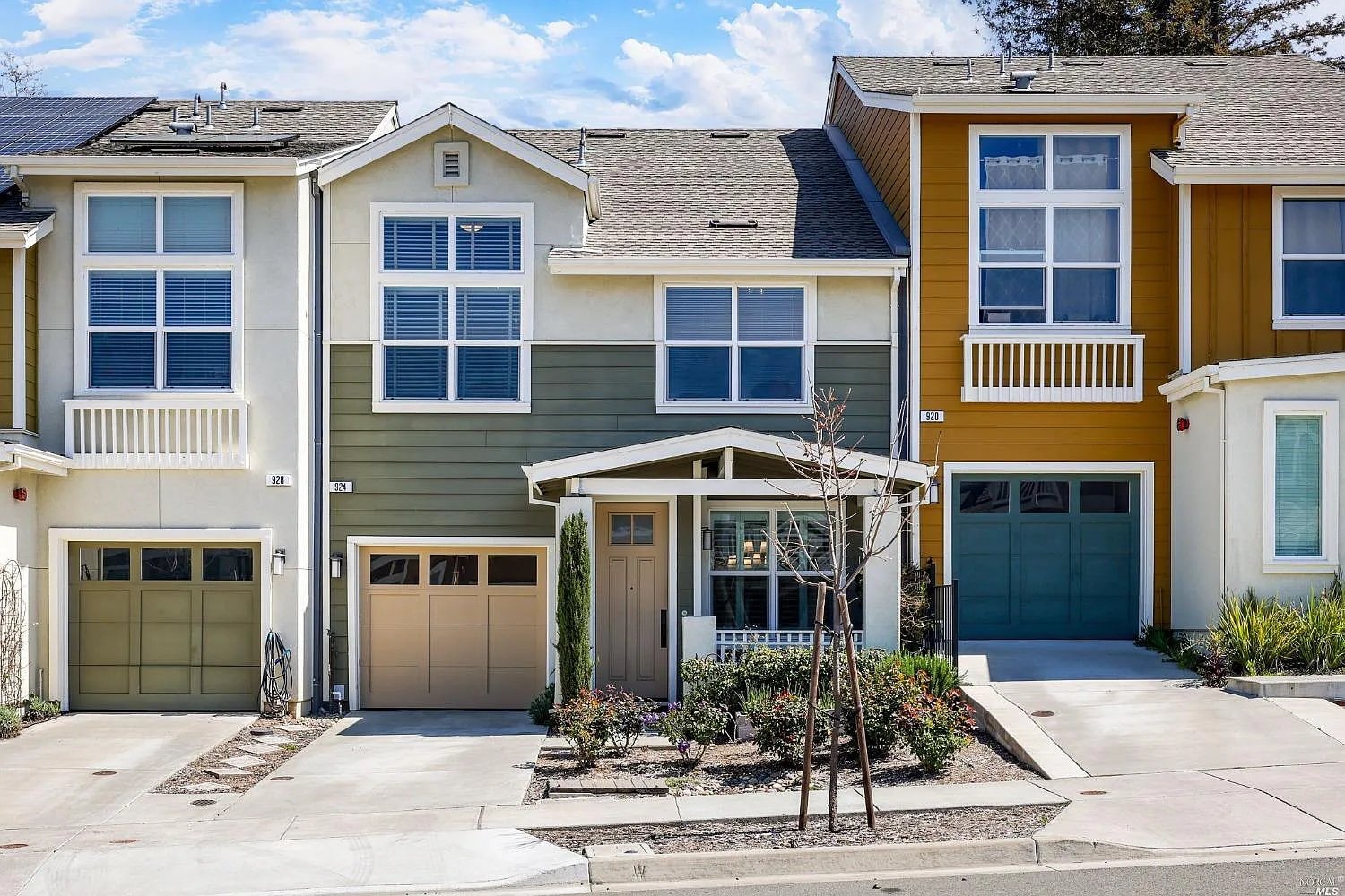 Modern townhouse exterior with green and yellow siding, large windows, and garages. Minimal landscaping with young trees and shrubs.