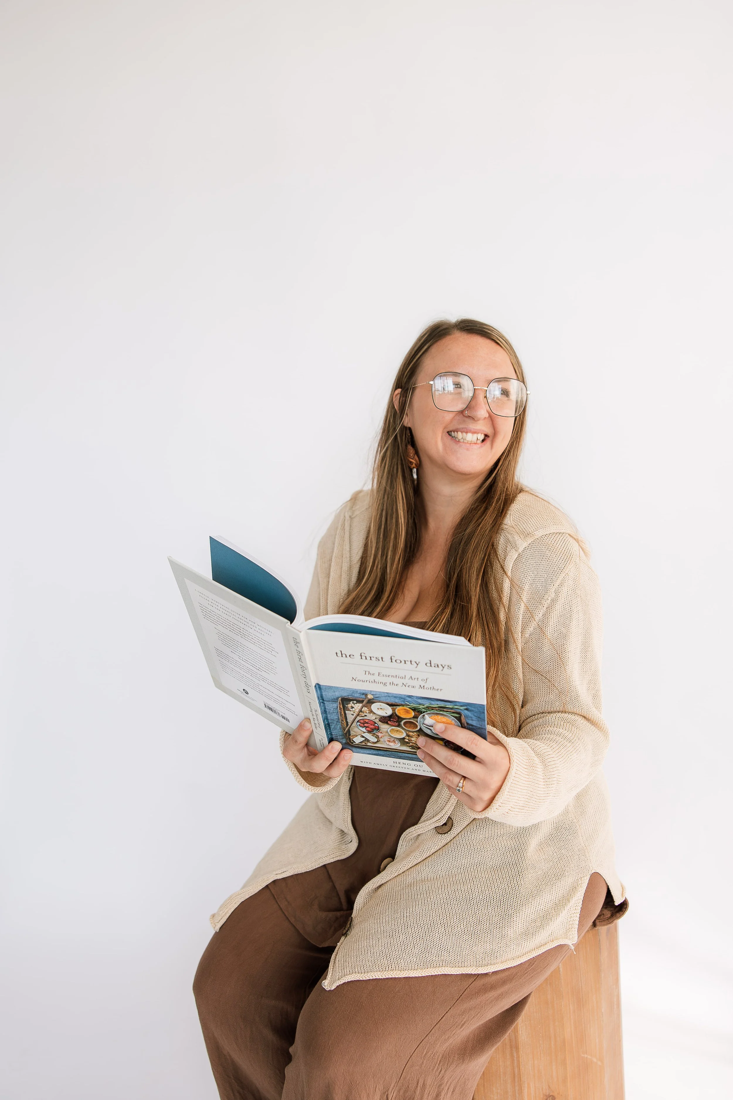 A birth doula with glasses reading a book titled "The First Forty Days" while sitting on a wooden stool against a plain white background.