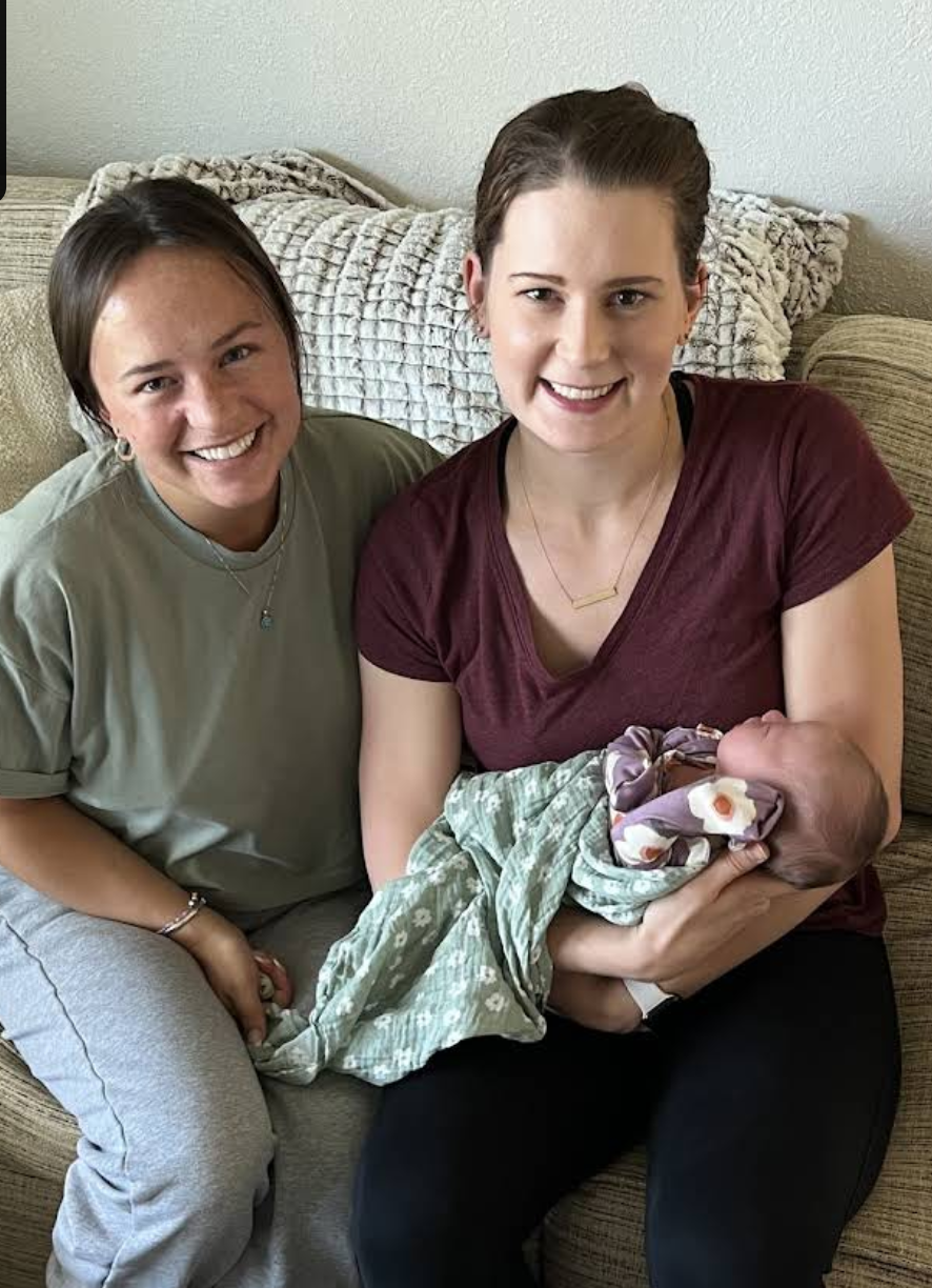 Home birth midwife and past client sitting on a beige couch, the midwife holding a newborn baby wrapped in a blanket.