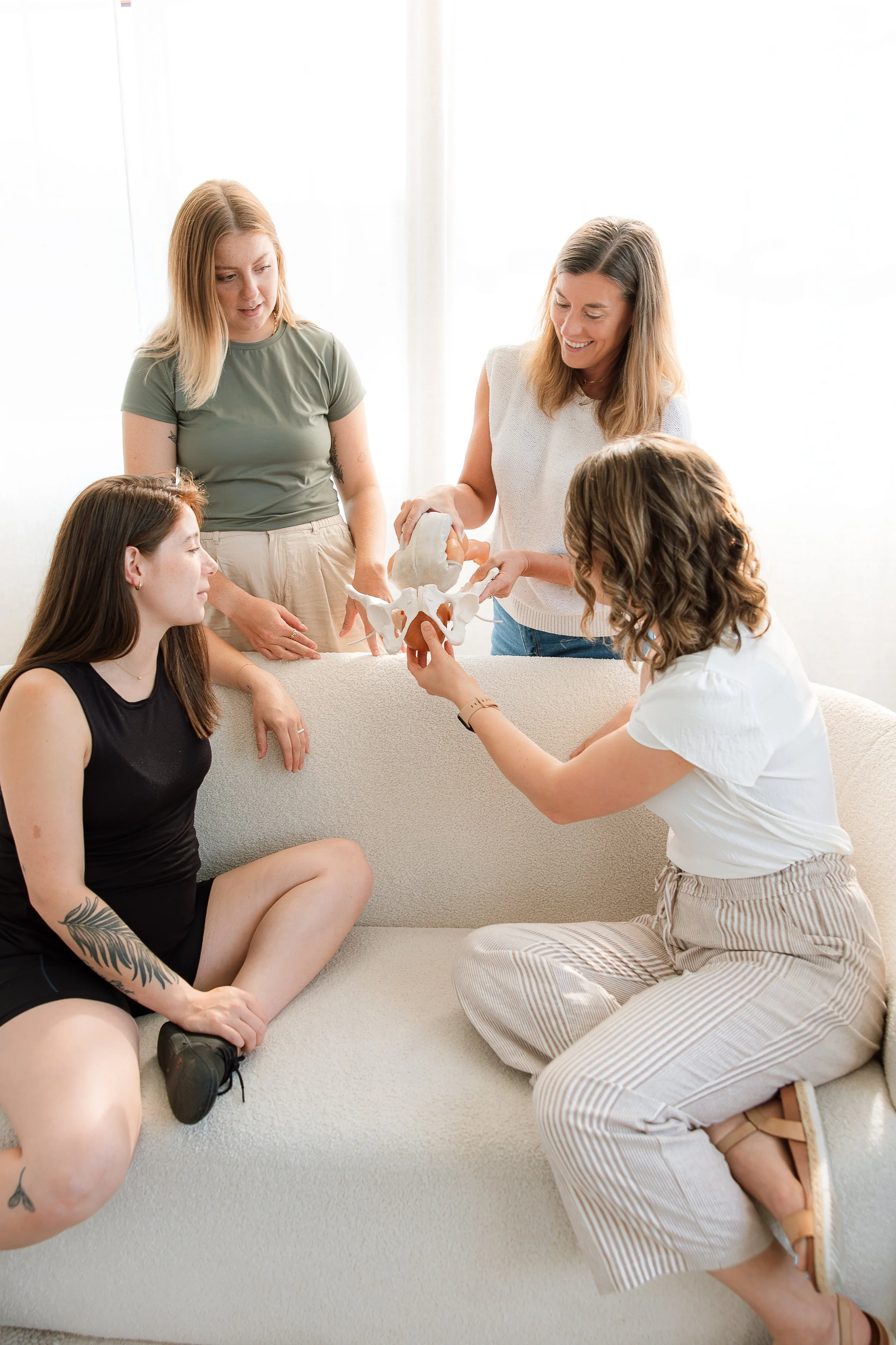 A group of birth doulas in a bright room, sitting and standing around a sofa, educating about labor support using a pelvis model.