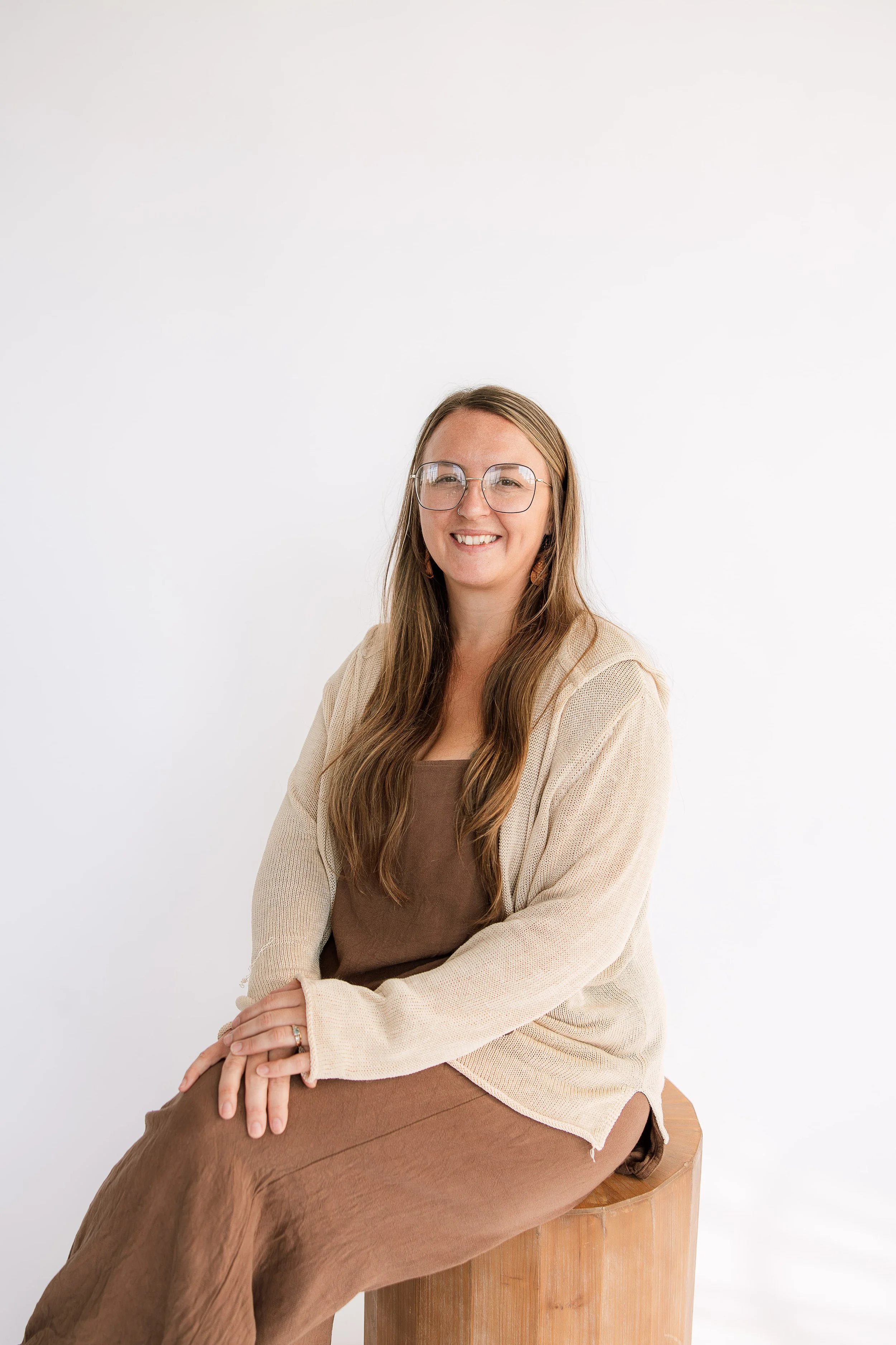 A birth doula with long brown hair, wearing glasses, a tan cardigan, and a brown dress, sitting on a wooden stool against a white background, smiling at the camera.