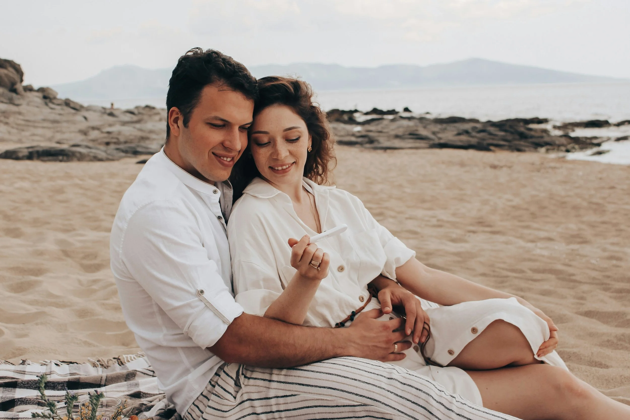 Husband and wife sitting on the beach excited and wondering what to do after a positive pregnancy test.