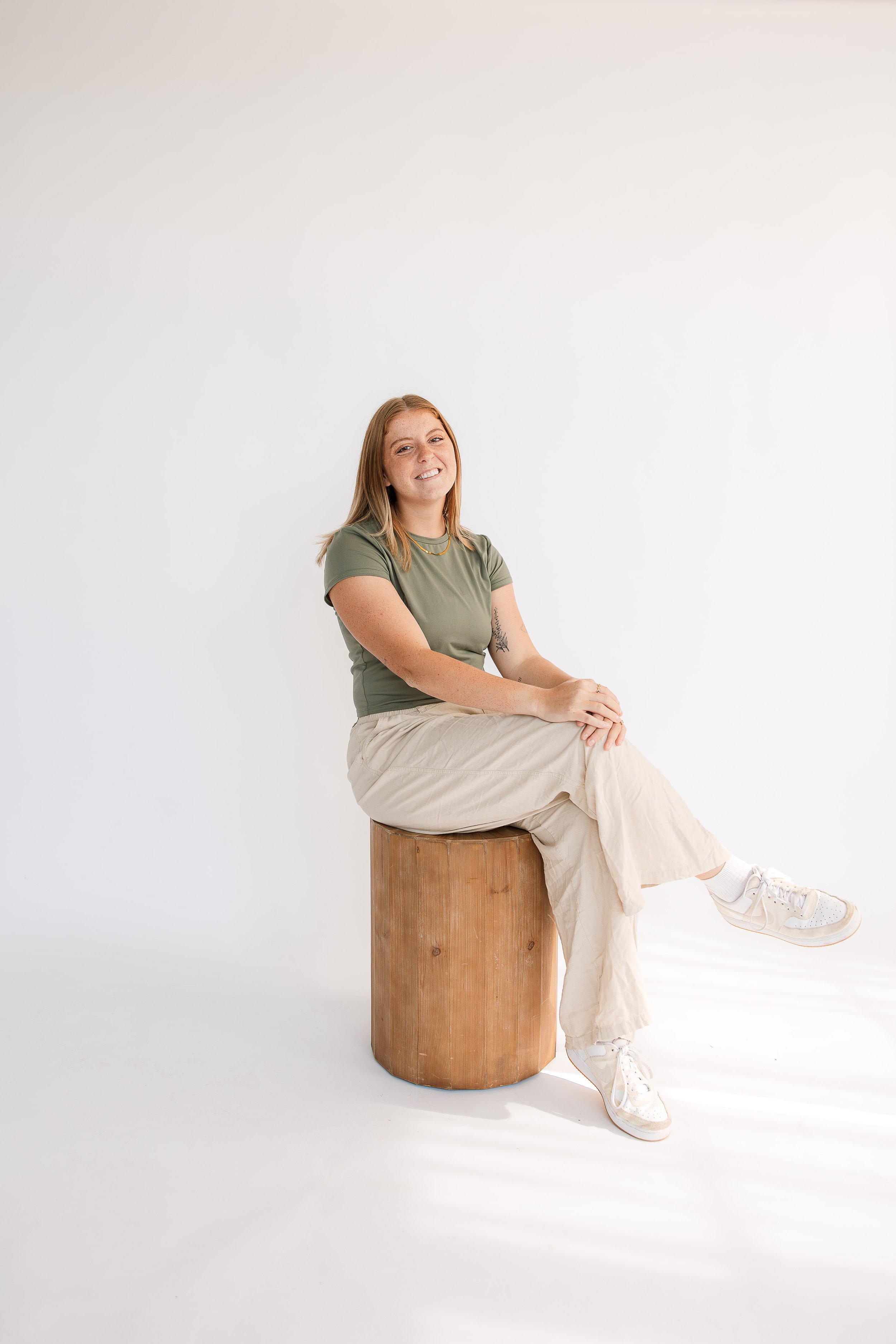 Birth doula in milwaukee wearing a green t-shirt and beige pants, sitting on a wooden stool against a plain white background, smiling.