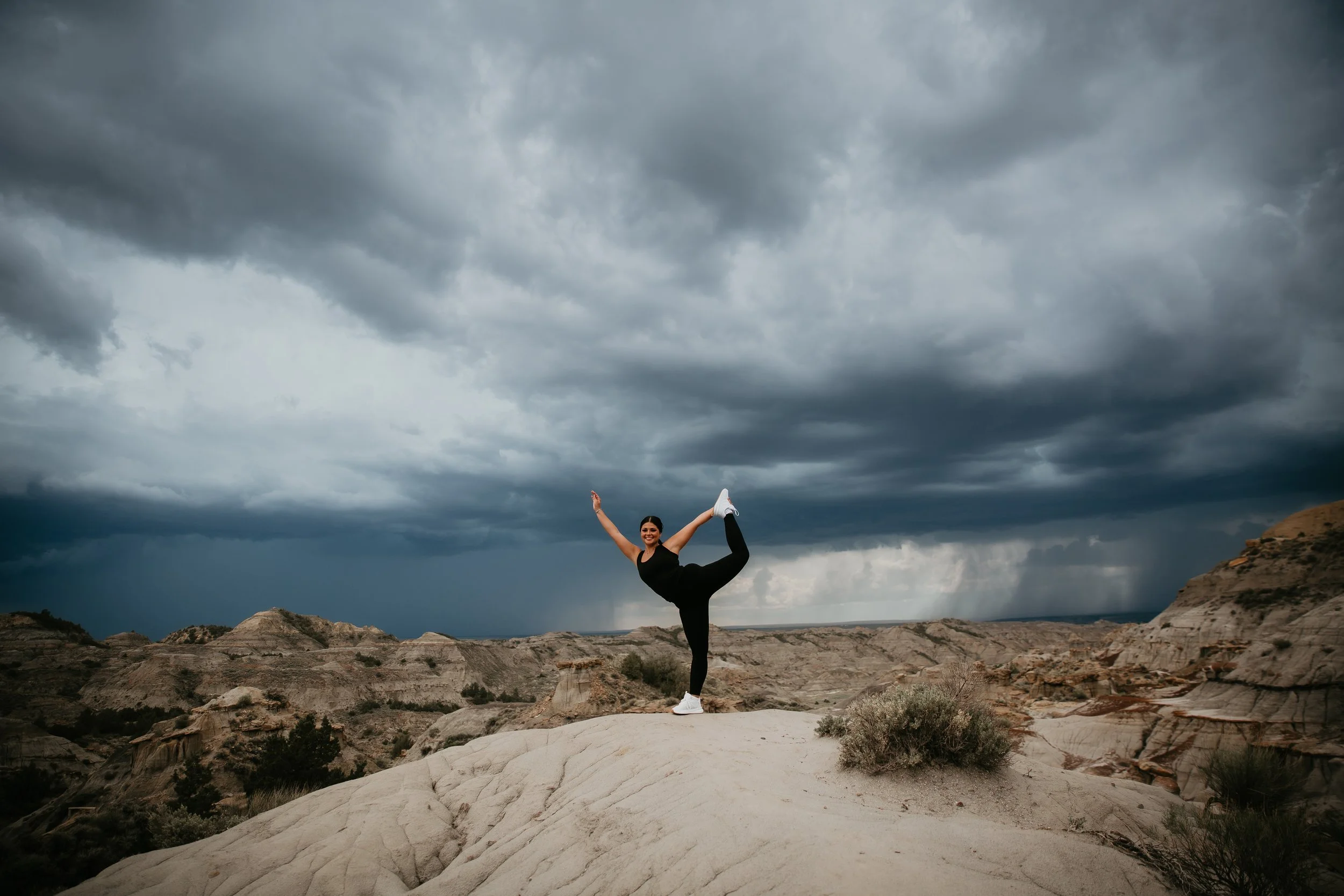 A woman in black athletic clothing performing a yoga pose on a rock formation outdoors, with dramatic cloudy skies overhead.