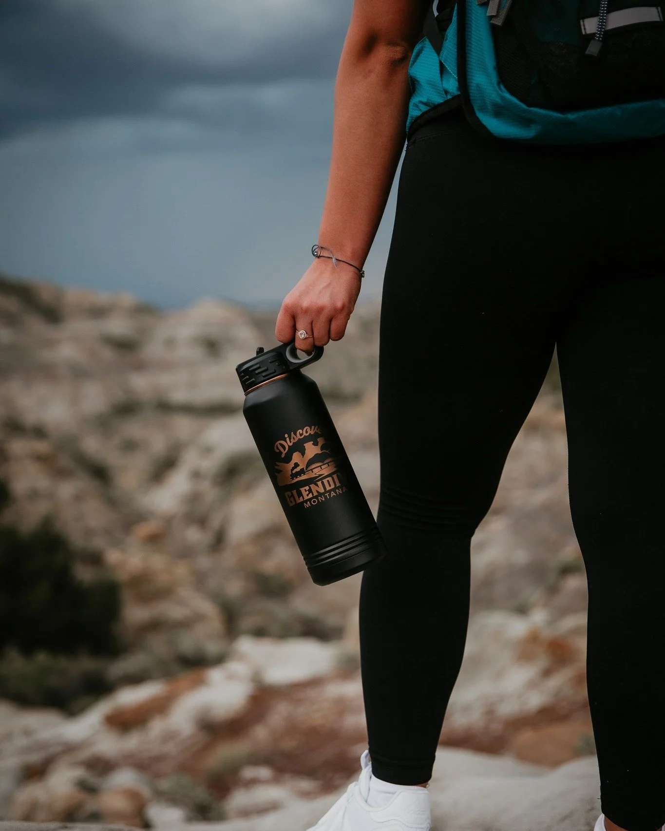 Person holding a black water bottle with 'Discover Glendive Montana' logo outdoors on rocky terrain, wearing black leggings, white sneakers, and a blue and black backpack.