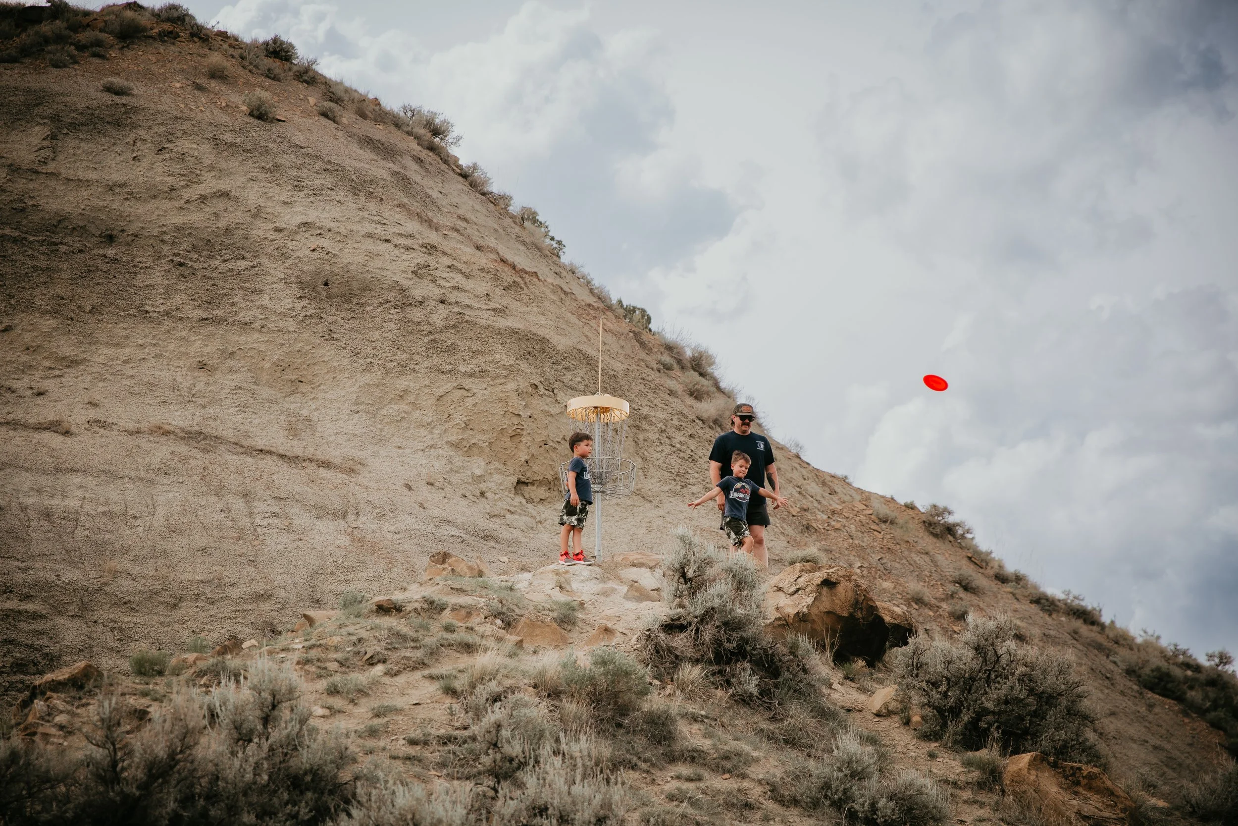 A man and two children flying a red disc golf disc in a desert-like area with rocky terrain and sparse vegetation, under a cloudy sky.